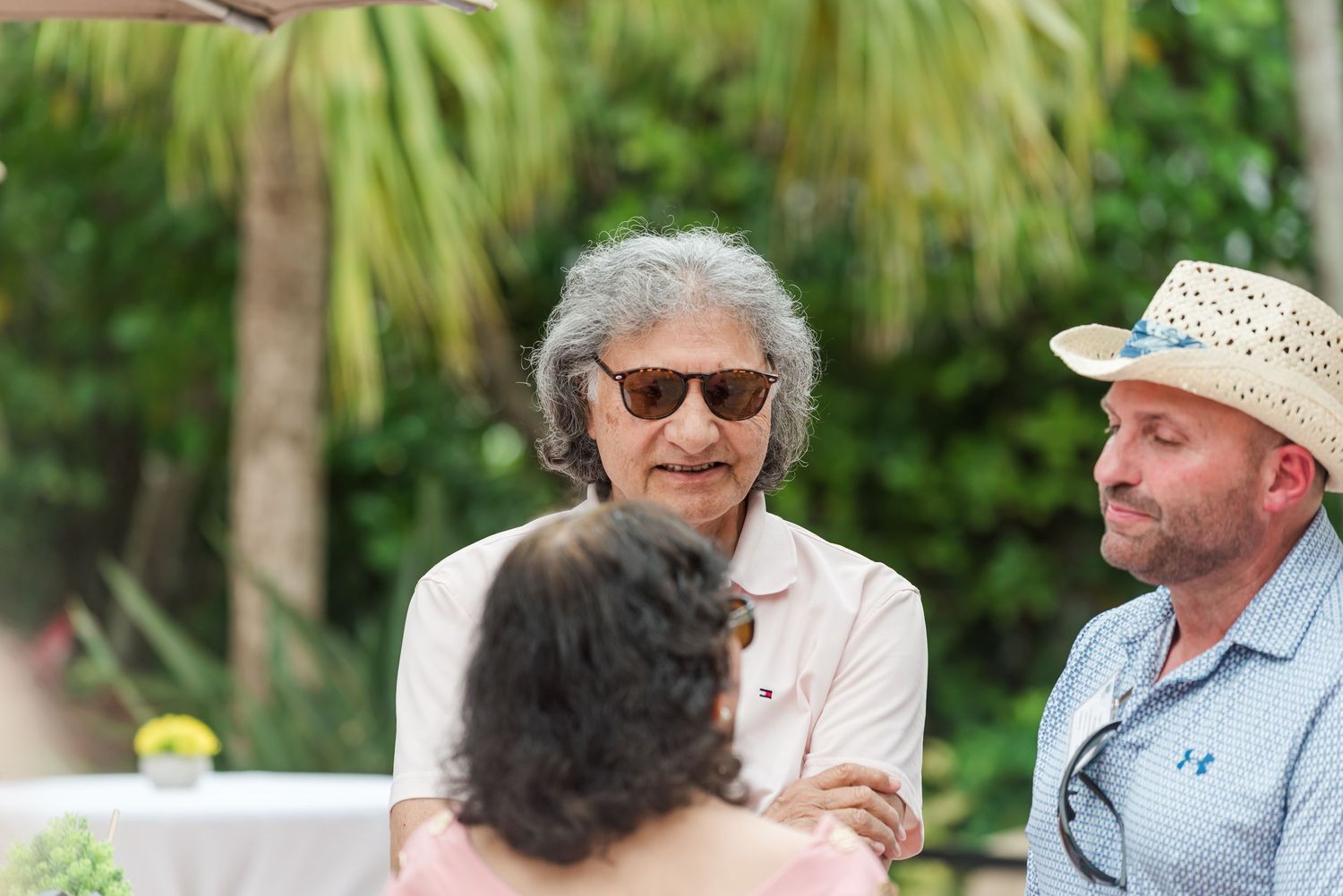 Three people engaged in conversation at a lush tropical outdoor gathering on a sunny day.