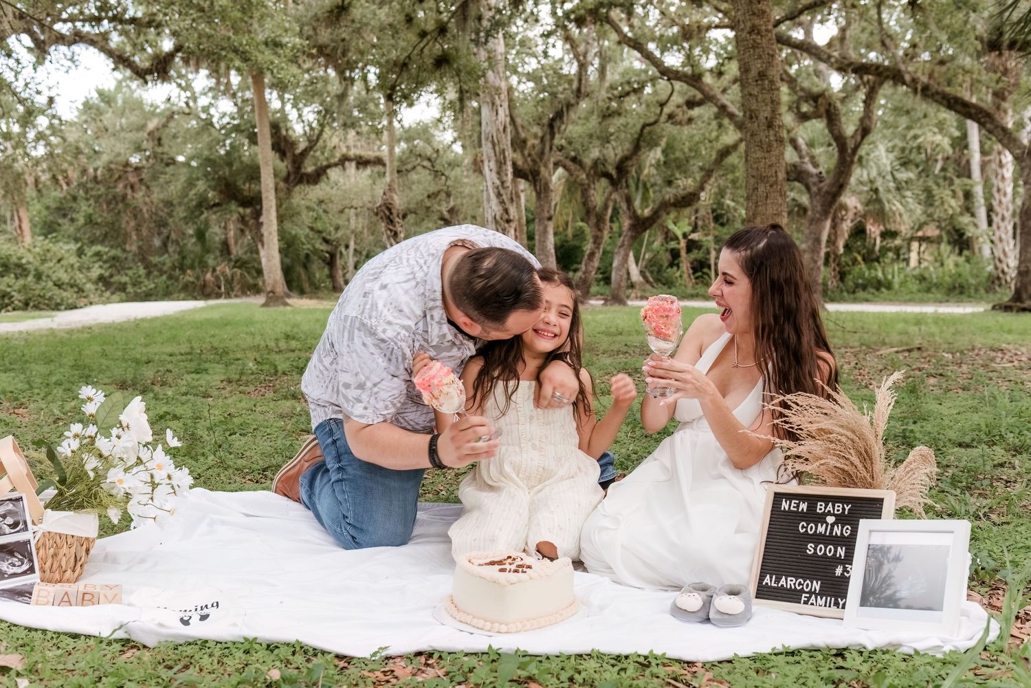 A romantic picnic scene in a park with a white blanket, snacks, and drinks spread out on the grass under trees.