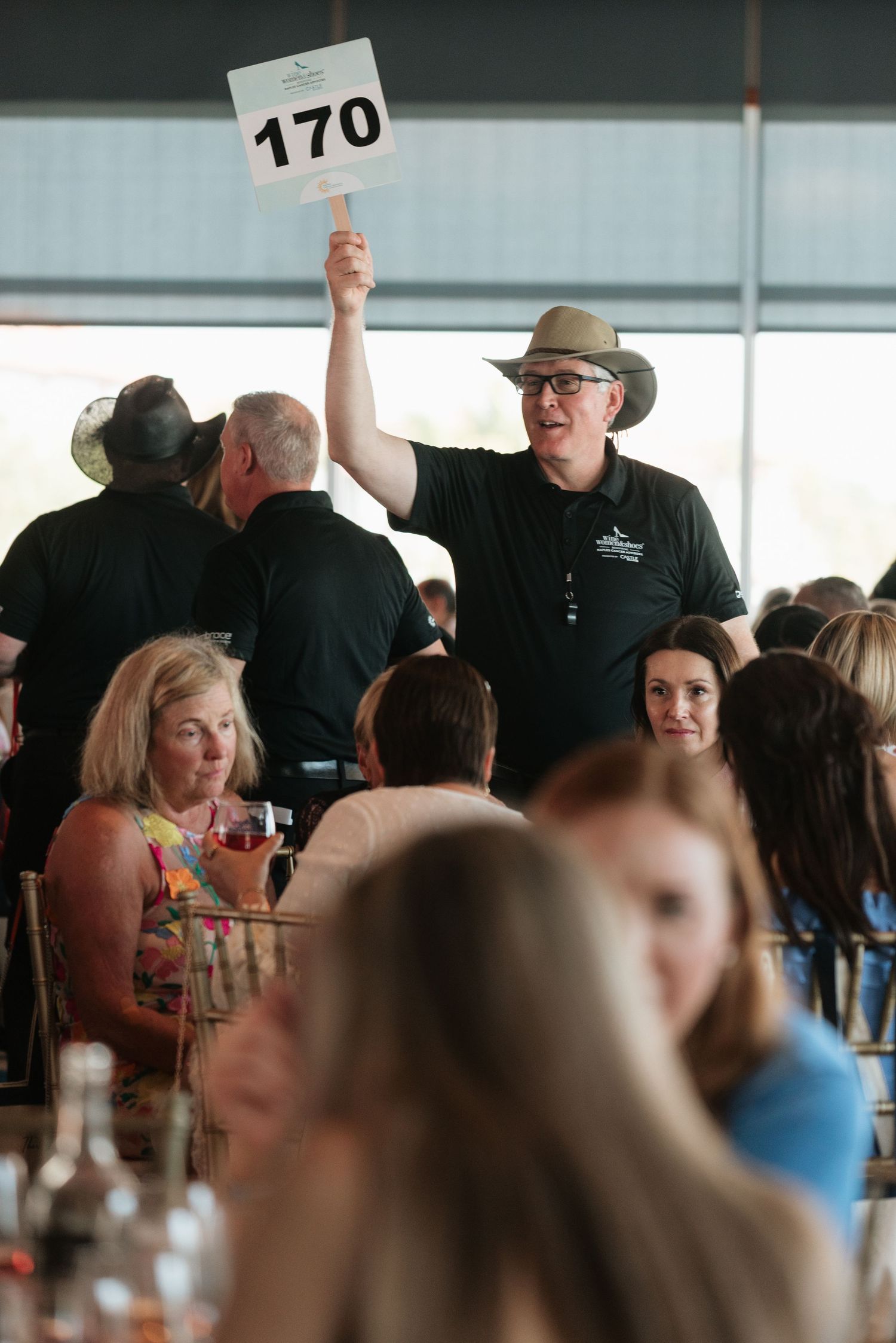 An auctioneer in a dark shirt and hat holds up bidding number 170 during a crowded indoor auction event.
