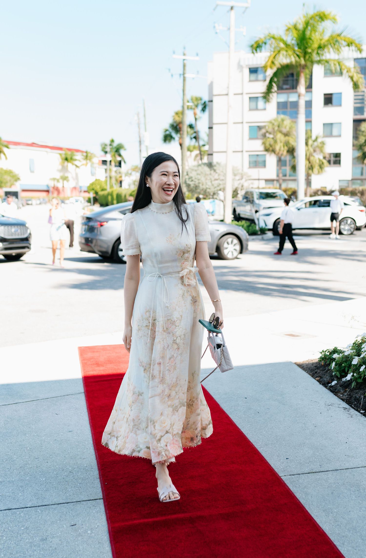 A woman in a flowing cream-colored dress walks down a red carpet outside on a sunny day.