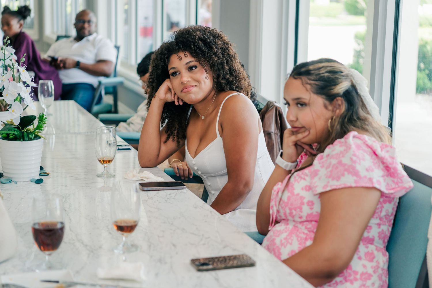 Two women sitting at a white bar table with wine glasses, smiling at the camera in a bright modern restaurant.