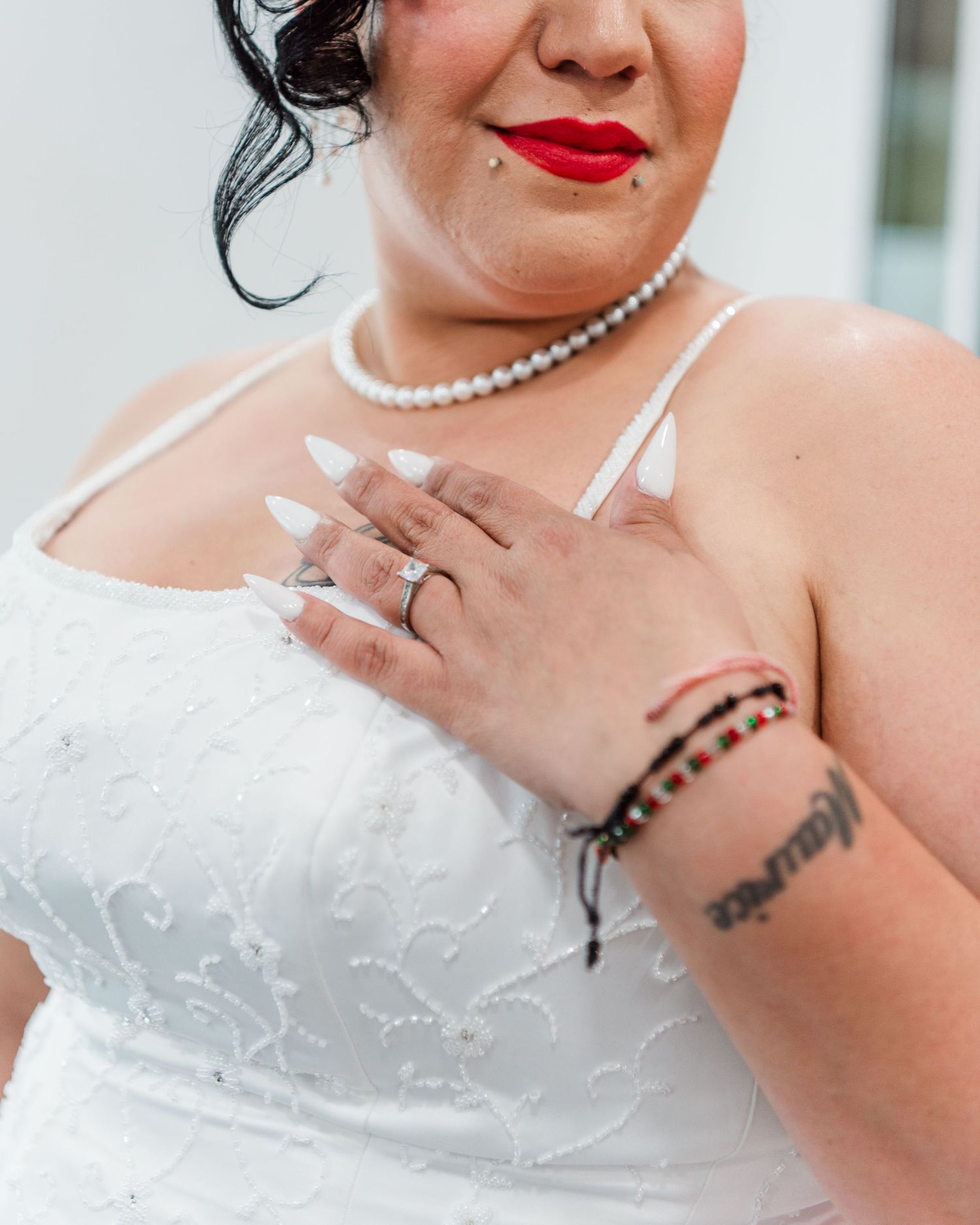 Close-up of a bride in white dress with pearl necklace and dark bracelet, showing wedding ring and lace details.
