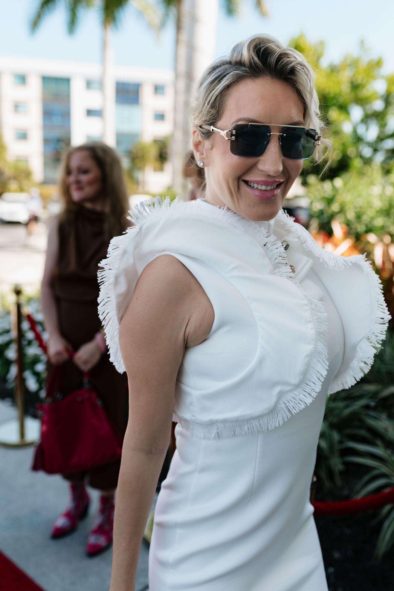 Person in white ruffled top and sunglasses smiles while posing outdoors near buildings and greenery.