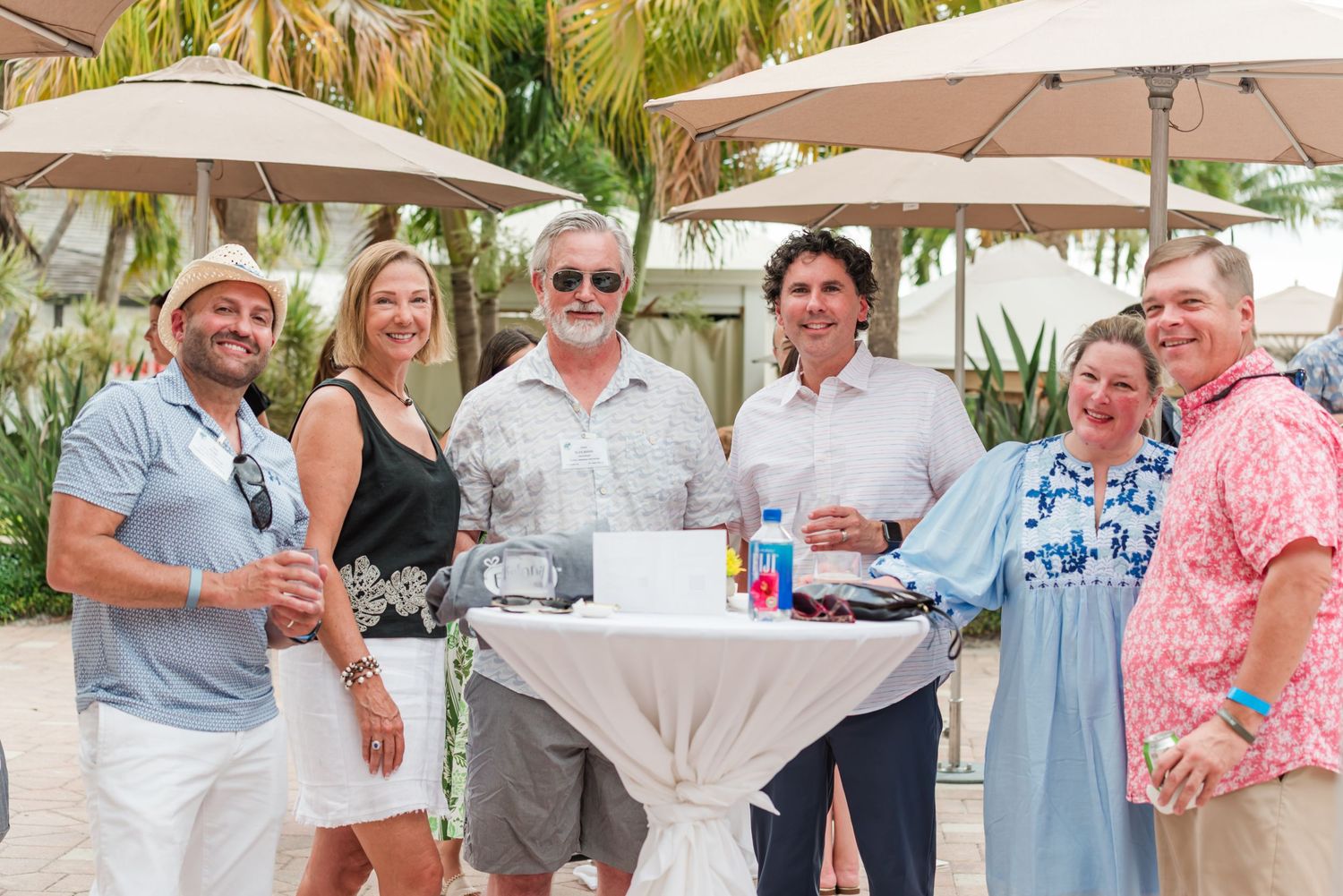 Group of six adults smiling together around a white cocktail table at an outdoor resort event.
