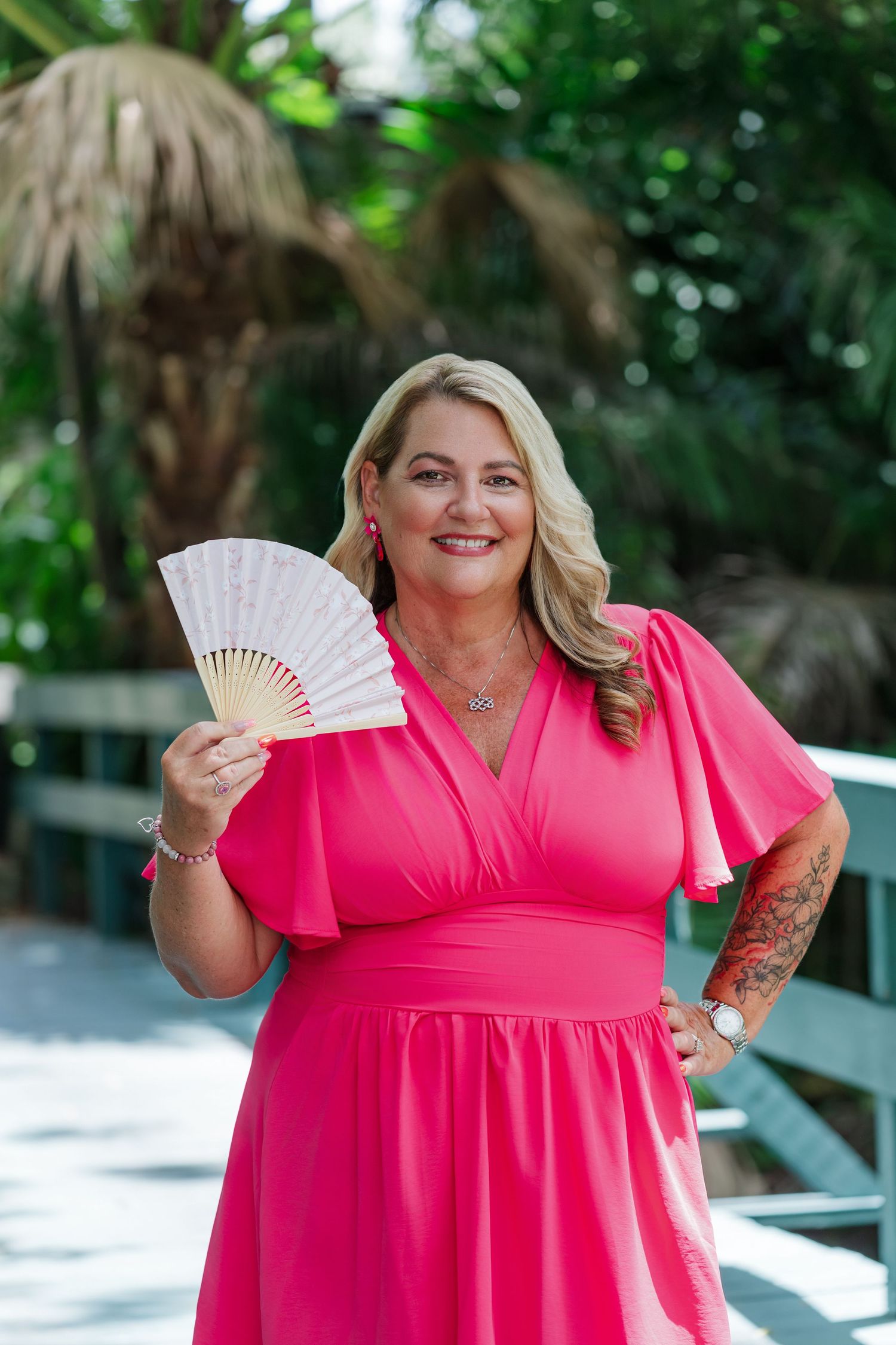 Smiling blonde woman in pink dress holding decorative fan outdoors among tropical greenery.