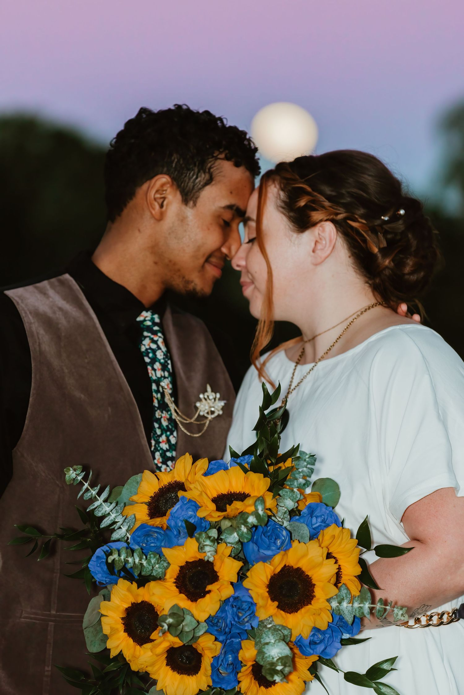 Romantic wedding couple shares tender moment with vibrant sunflower and blue flower bouquet at sunset.