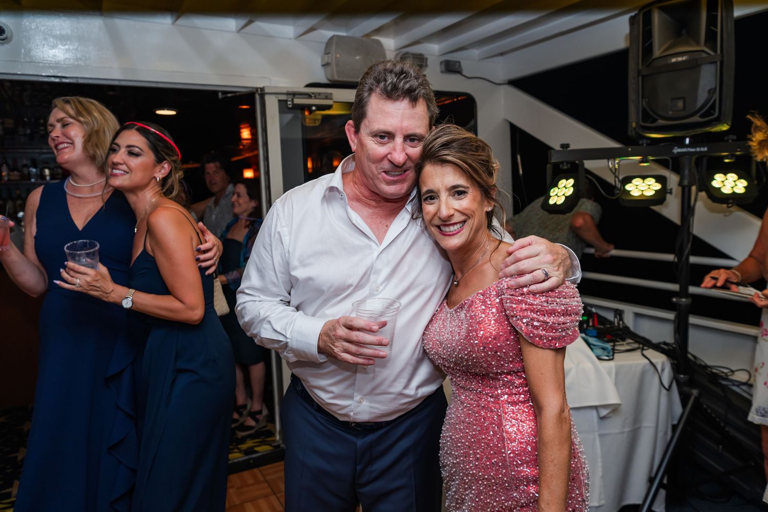 Couple posing together at an elegant evening party, woman in pink sequin dress, man in white shirt, guests dancing behind.