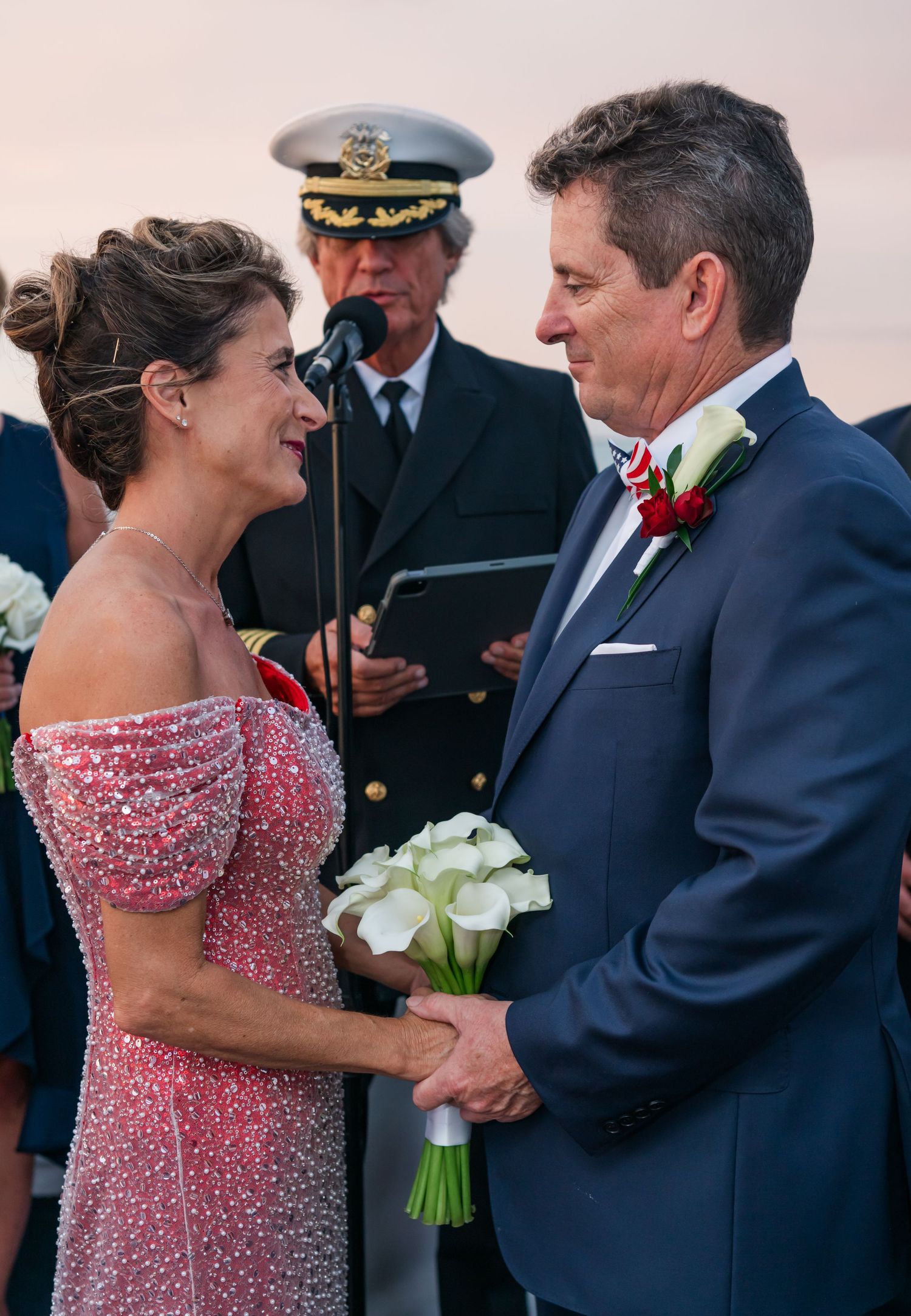 Couple exchanging vows at outdoor wedding ceremony, bride in pink beaded gown holding white calla lily bouquet, officiant in naval uniform.