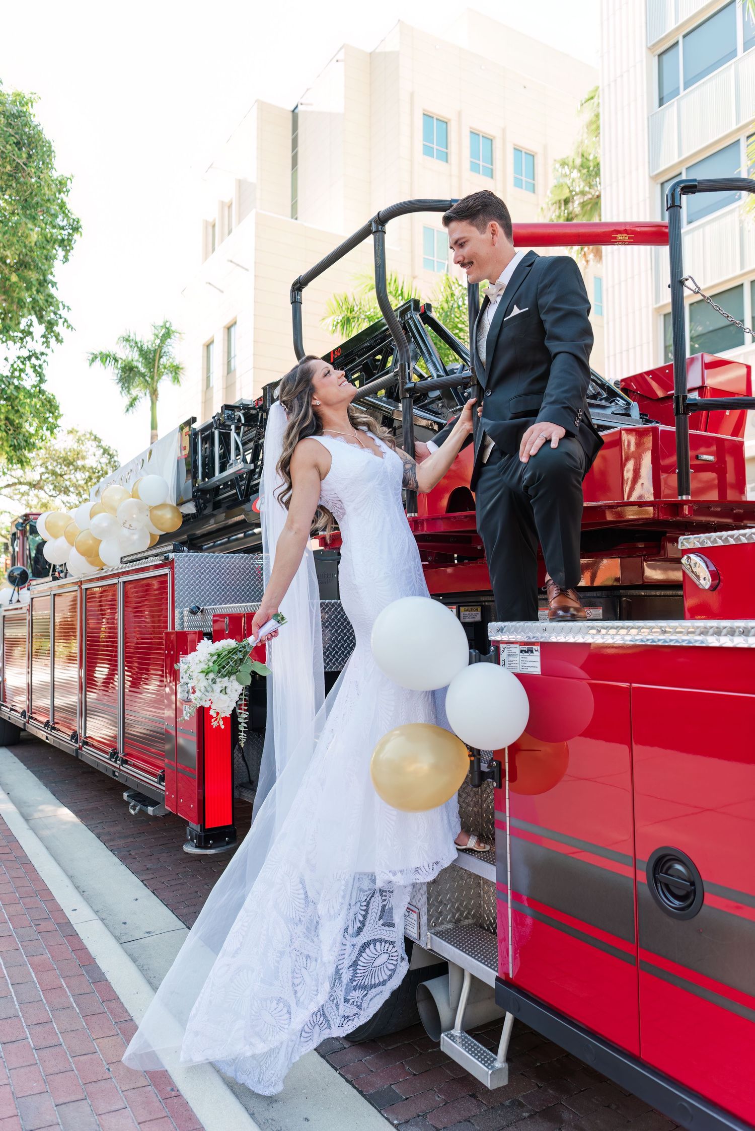 Bride and groom posing on a decorated red fire truck with gold and white balloons on their wedding day.