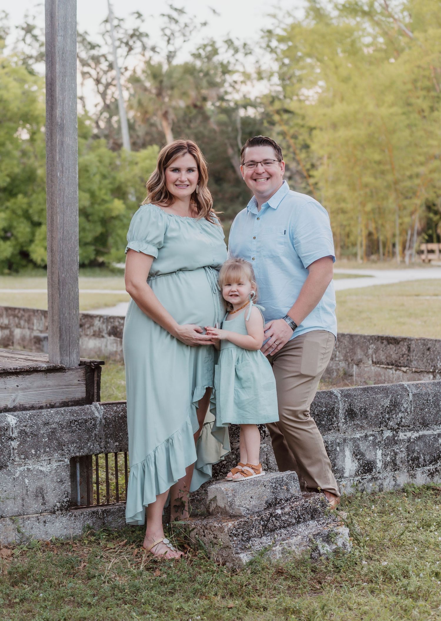 A family of three poses outdoors on stone steps wearing coordinating mint and blue outfits in a natural setting.