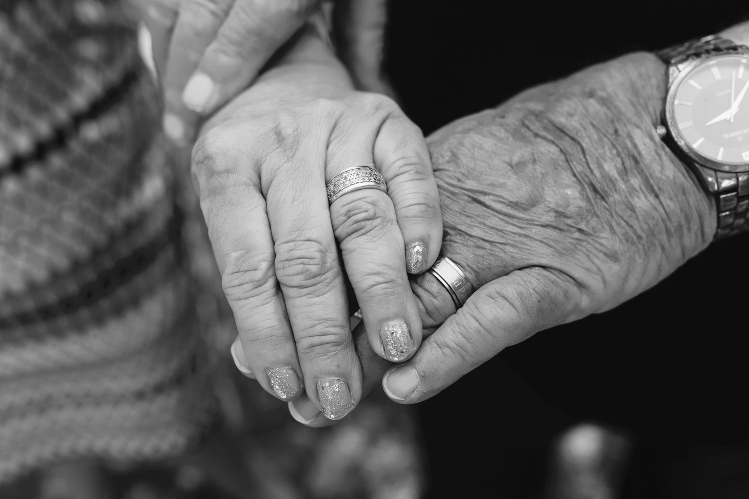 Black and white photo of elderly couple holding hands, showing wedding rings and a wristwatch, symbolizing lasting love.