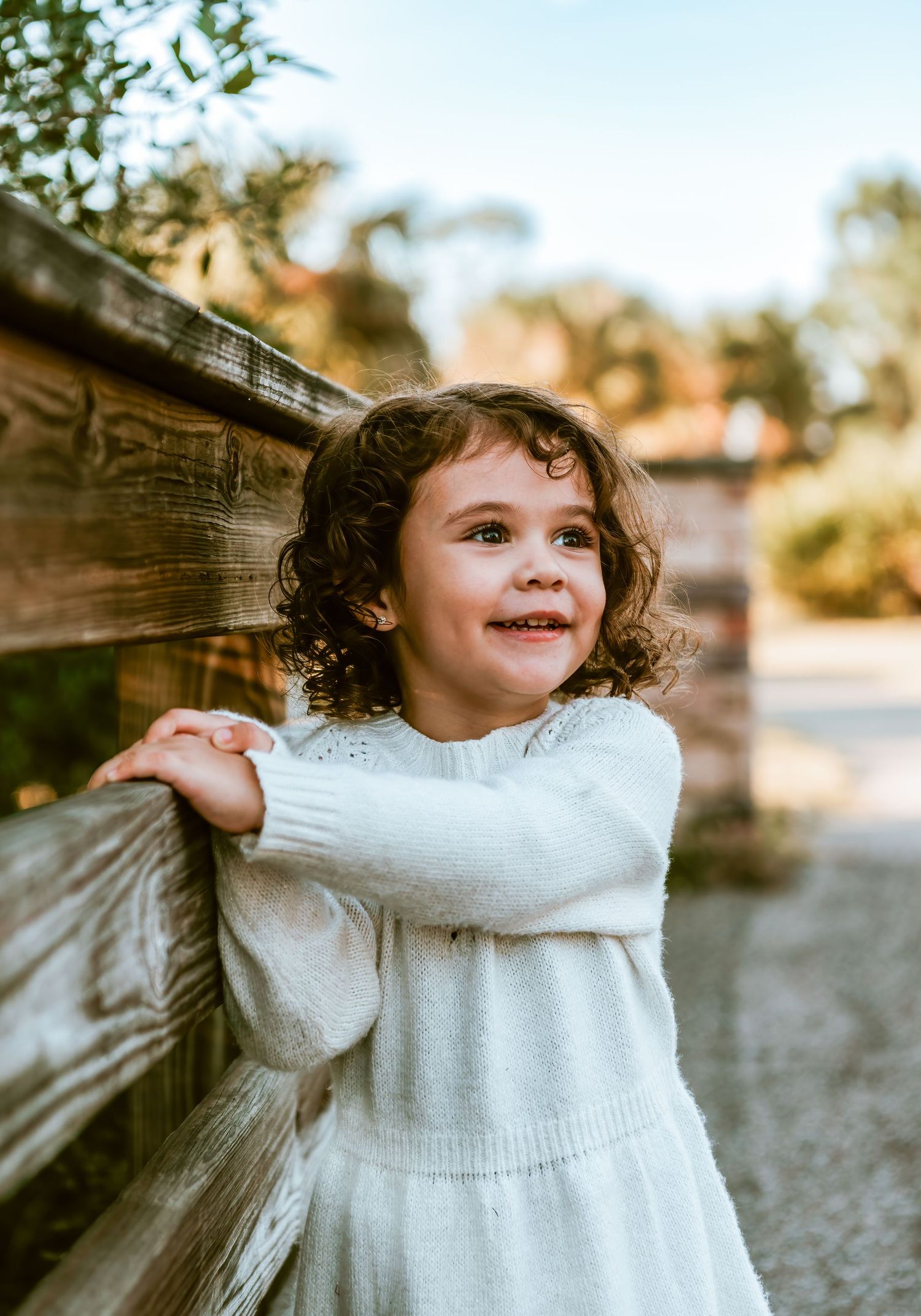 Young child in white sweater dress smiles while holding onto wooden railing outdoors in warm sunlight.