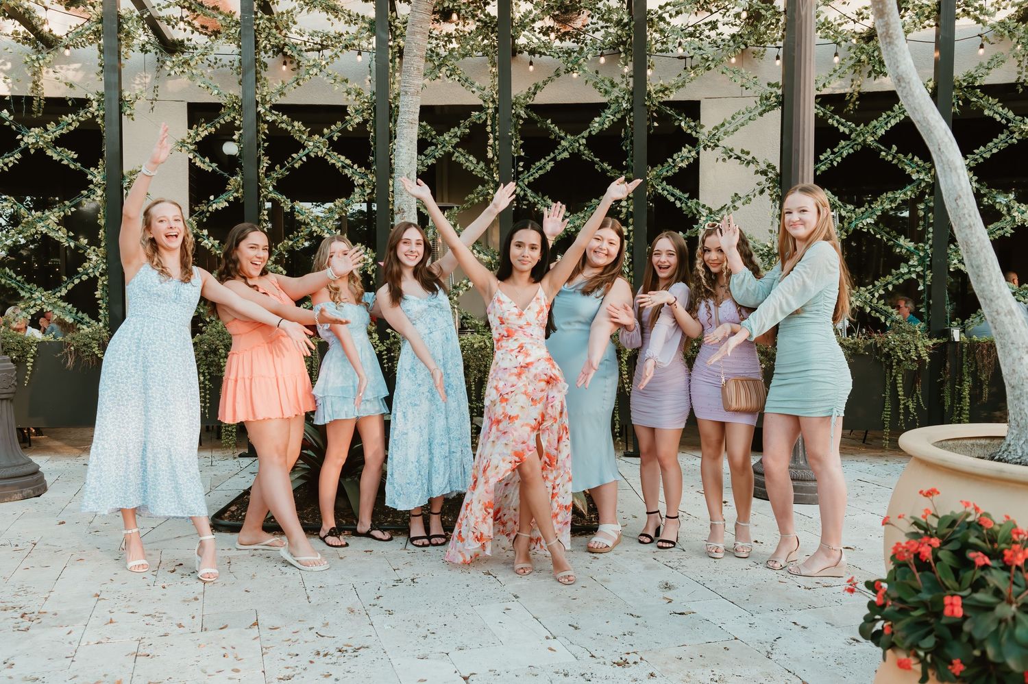 A group in pastel dresses celebrate joyfully under outdoor string lights at a garden party venue.