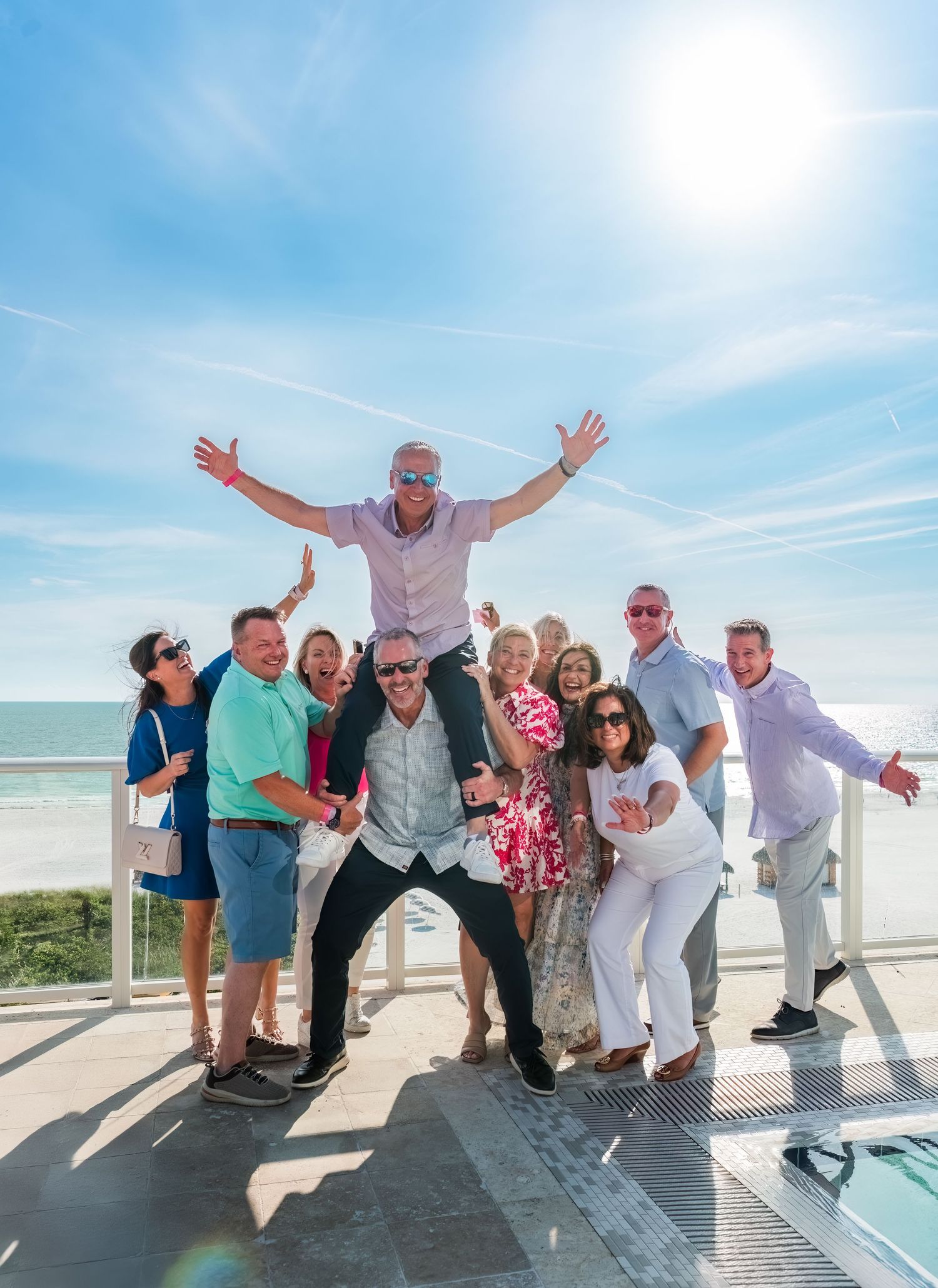Group of friends posing together at a beach boardwalk on a sunny day with someone jumping in celebration.
