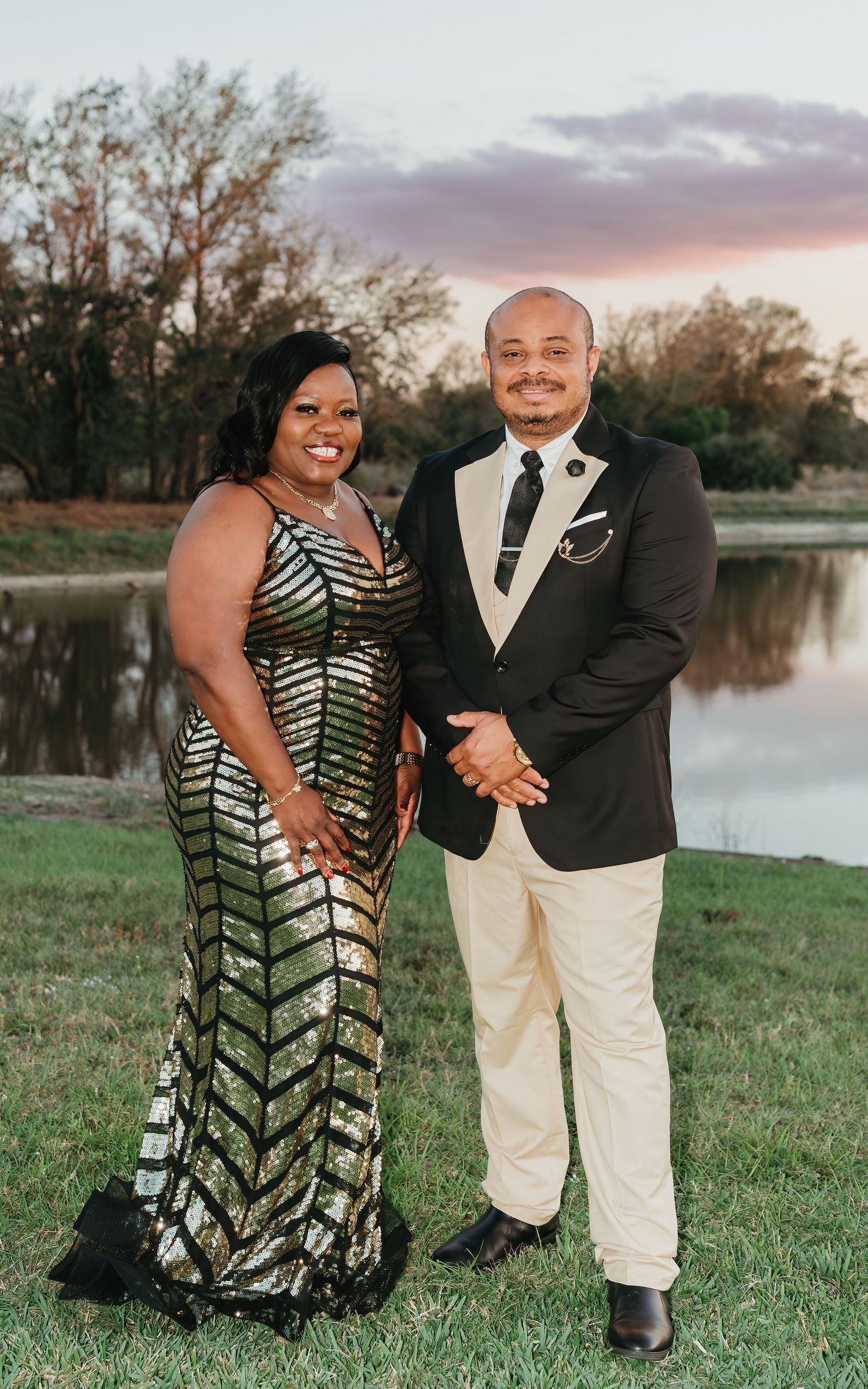 A couple poses by a serene lake at sunset while wearing formal attire including a sparkling dress and light-colored suit.