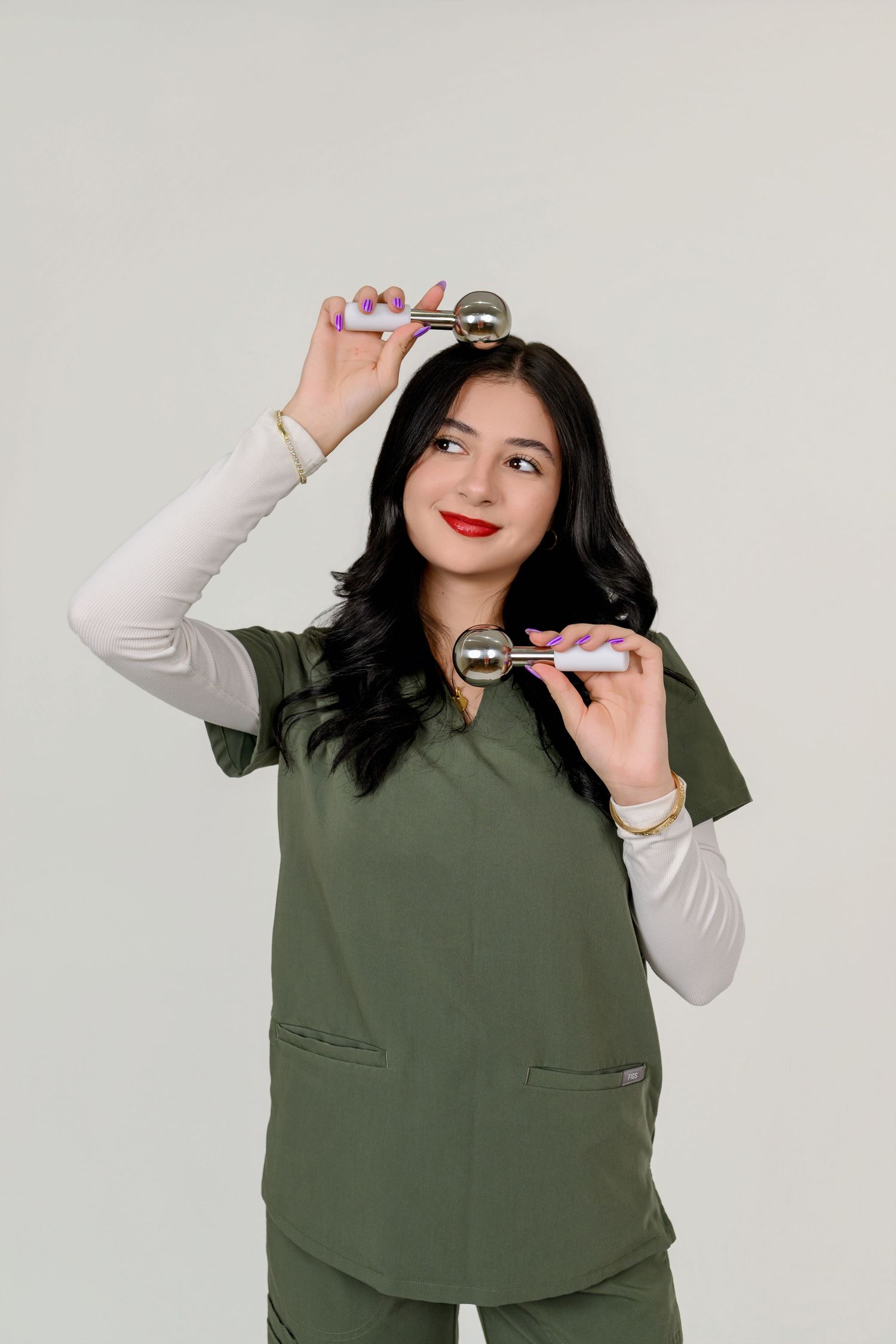 Female healthcare worker in olive green scrubs holding a facial roller massager tool against a white background.