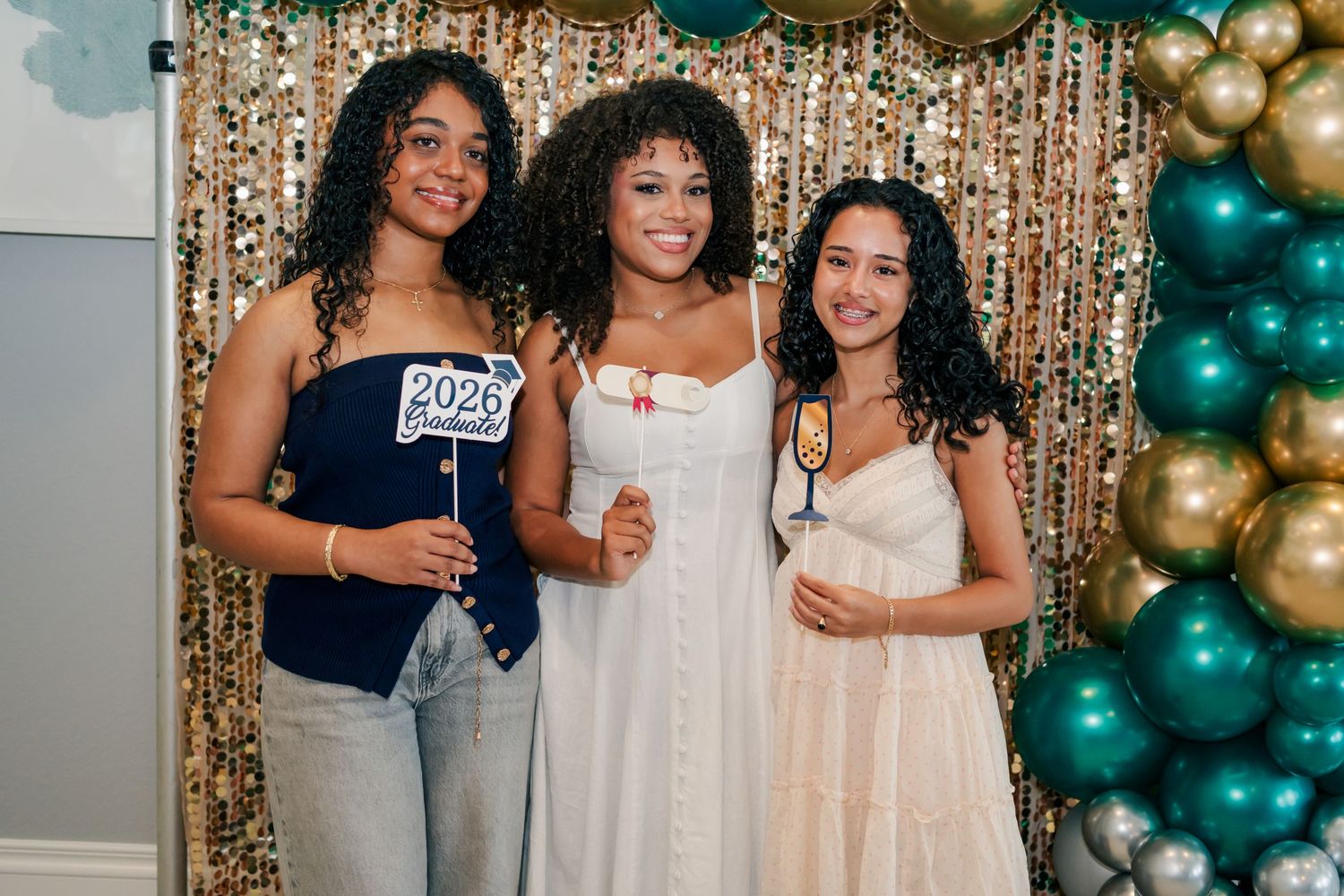 Three young women pose at a New Year 2026 party with gold and teal balloon decorations.