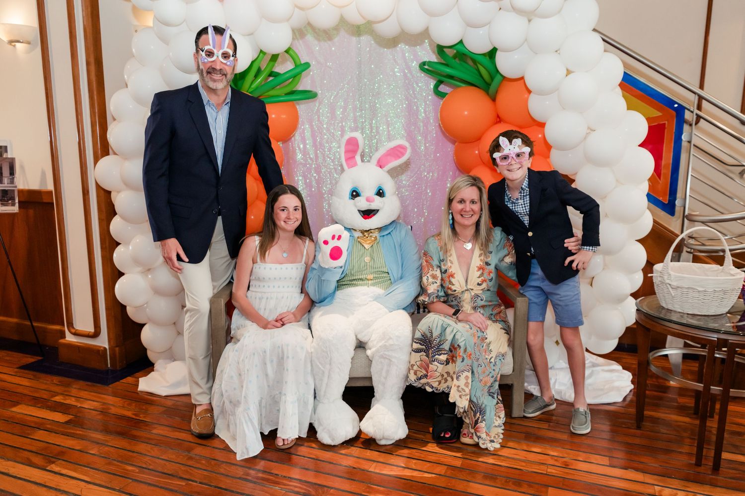 Group posing with Easter Bunny mascot under festive balloon arch decorated with carrots at Easter celebration.