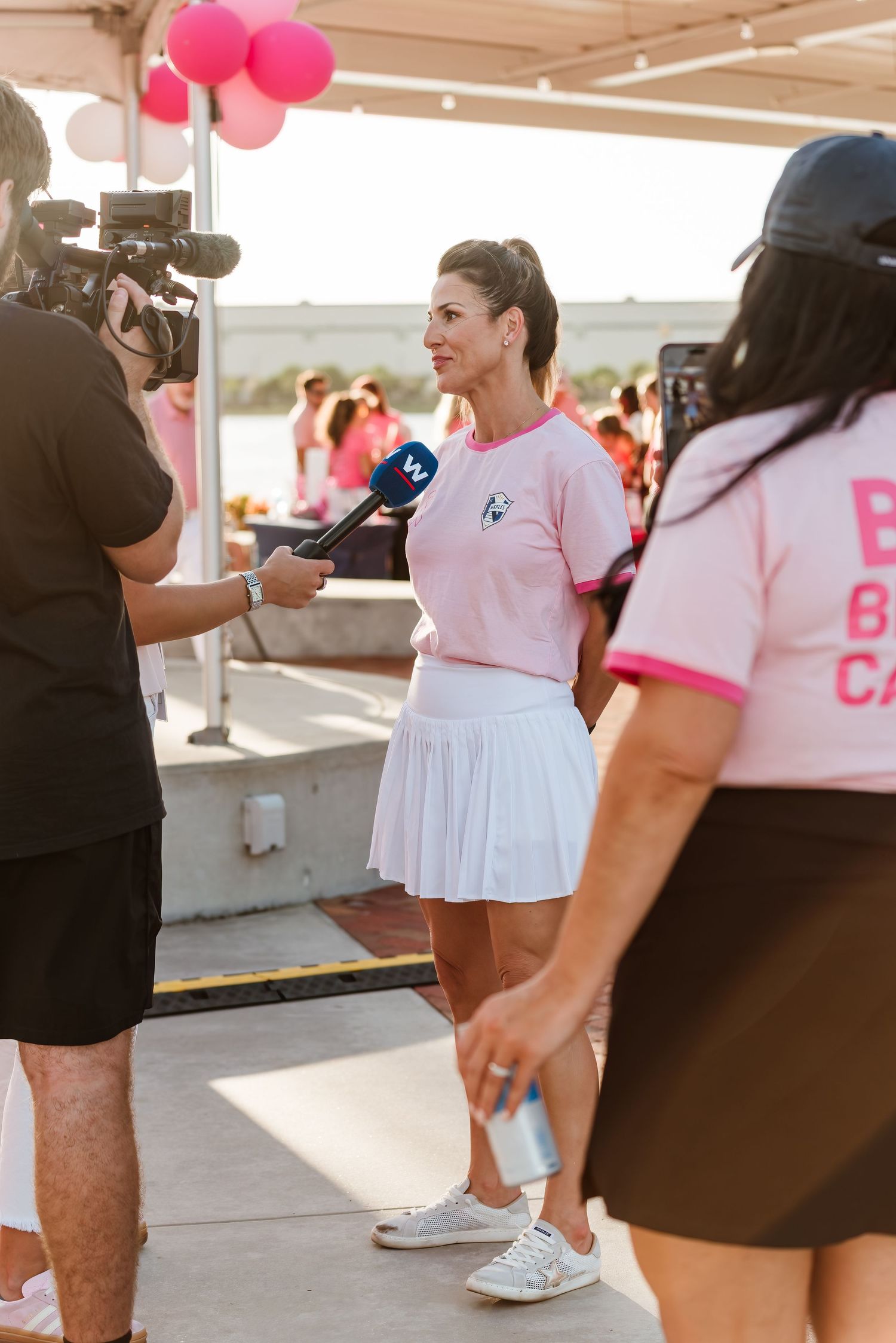 Camera crew interviews someone wearing a pink top and white tennis skirt at an outdoor event with pink balloons.