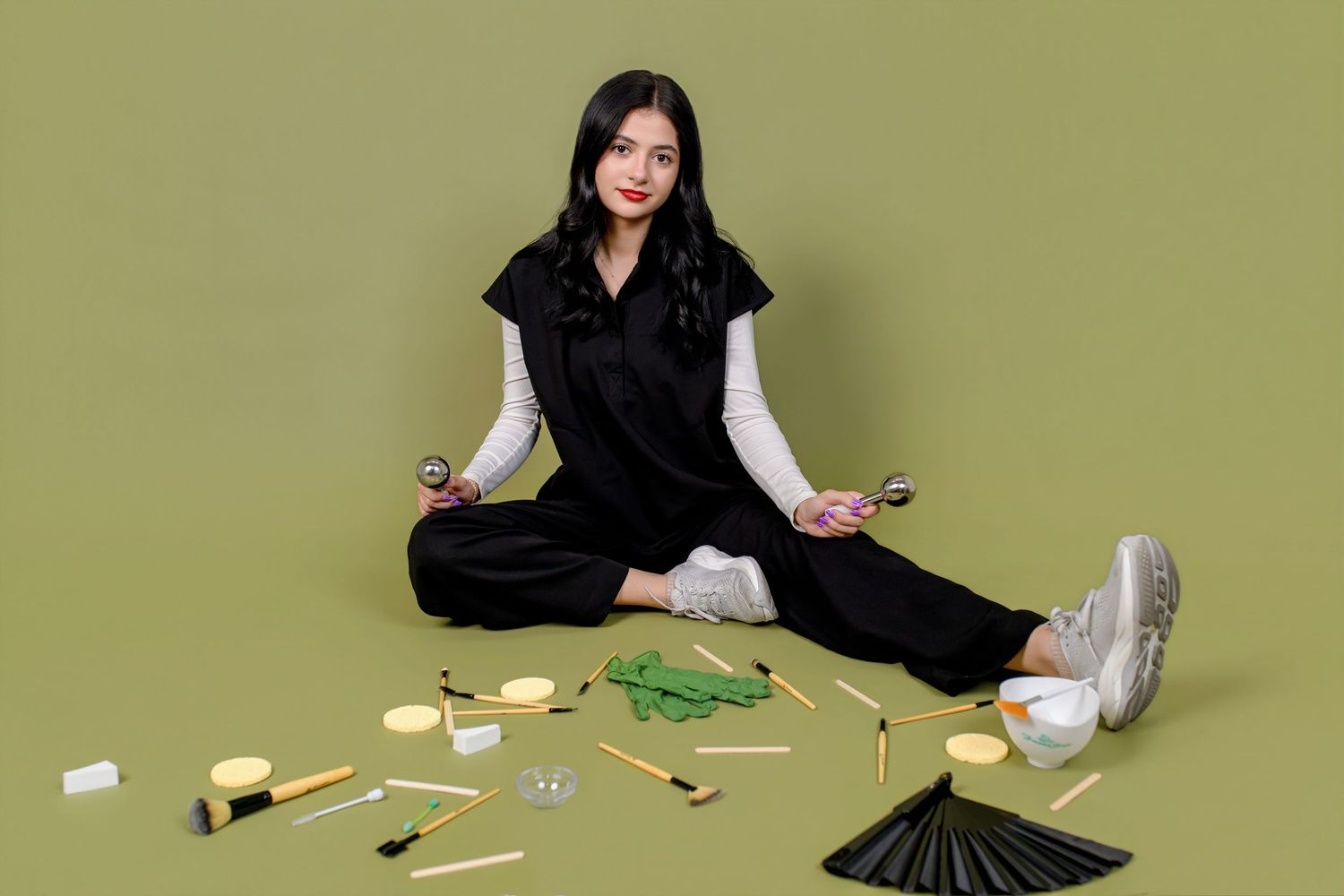 Woman in black outfit sits meditating surrounded by beauty tools on green background.