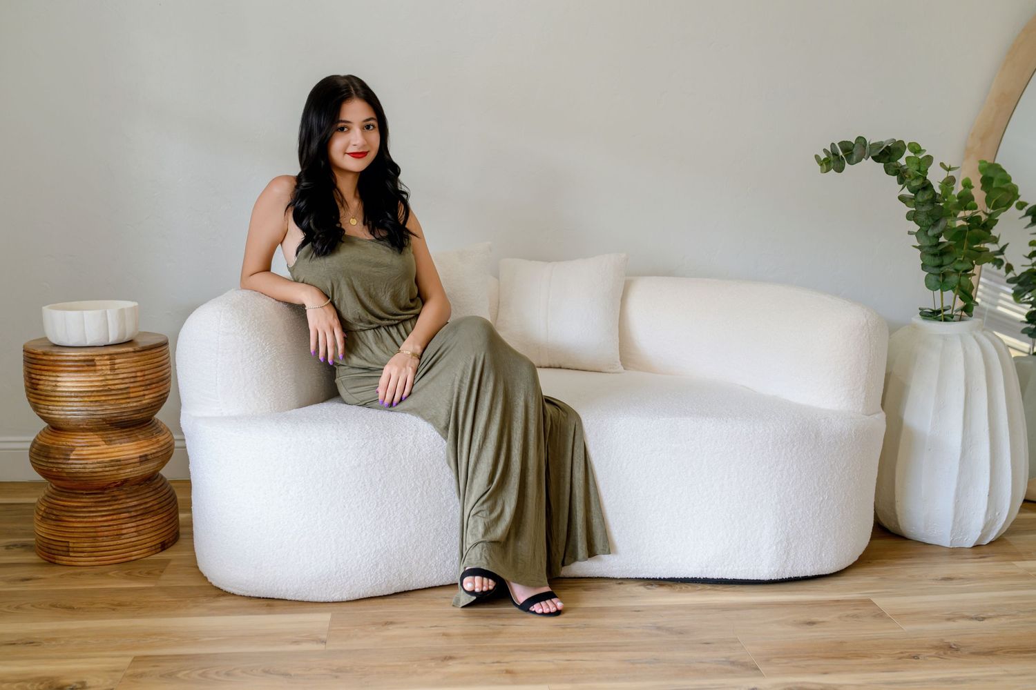 Woman in olive green maxi dress sitting on white boucle sofa with wood side table and plant decor.