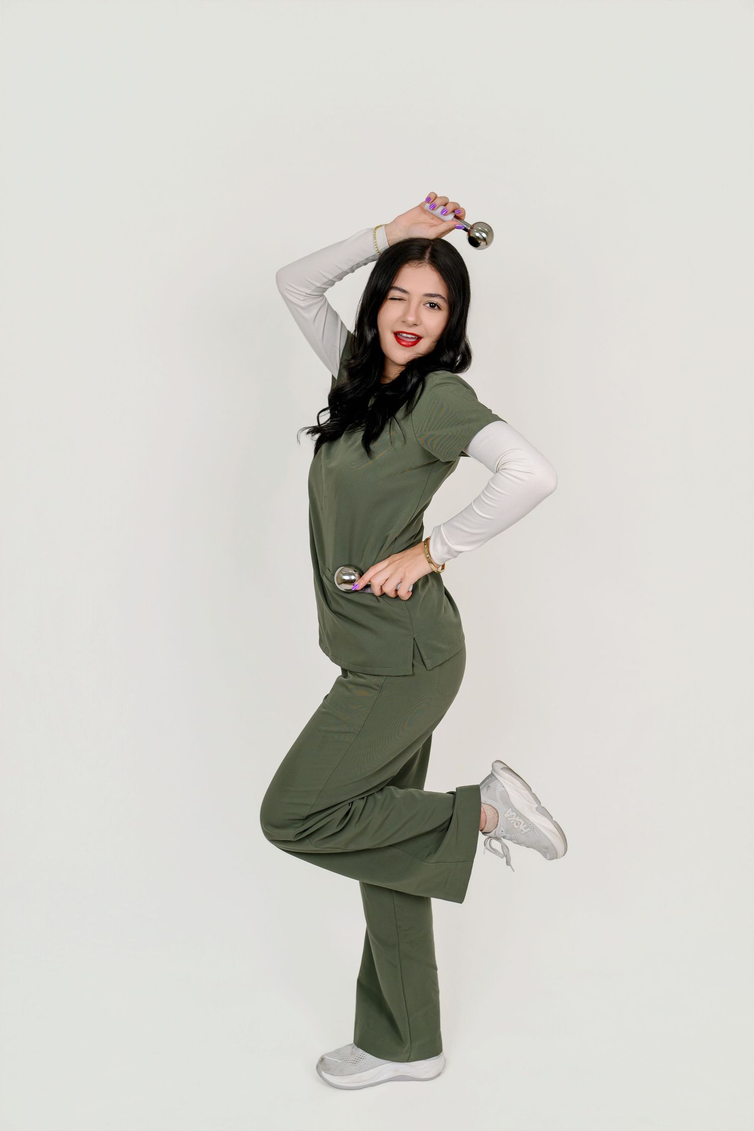 Woman in olive green medical scrubs poses playfully, holding stethoscope, wearing white sneakers on light background.