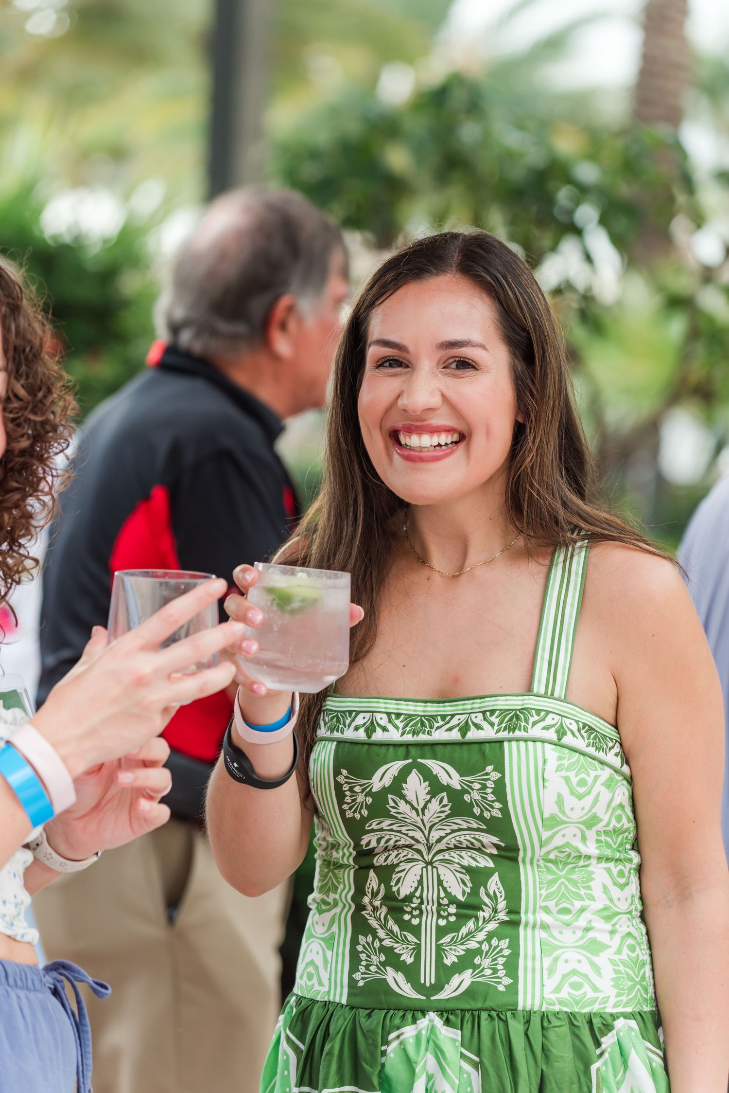 A smiling brunette woman in a green dress toasts drinks with someone at an outdoor social event.
