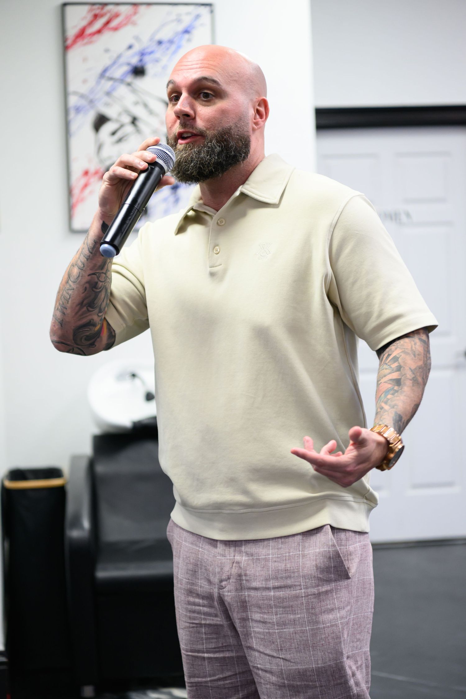 Tattooed bald man with beard speaking into microphone, wearing cream polo shirt and gray trousers indoors.