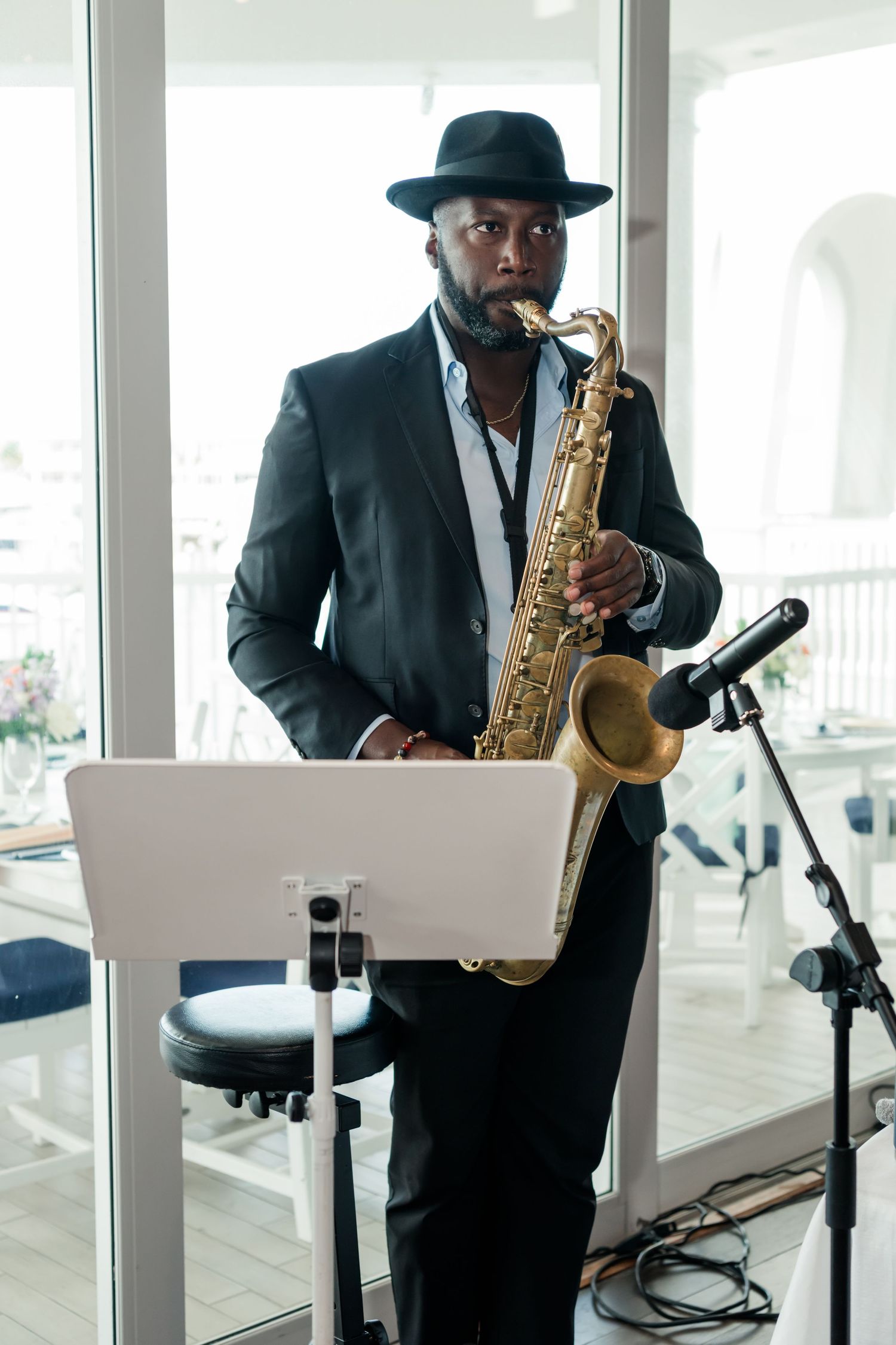 A jazz musician in a black suit and hat plays a golden saxophone at a music stand indoors.