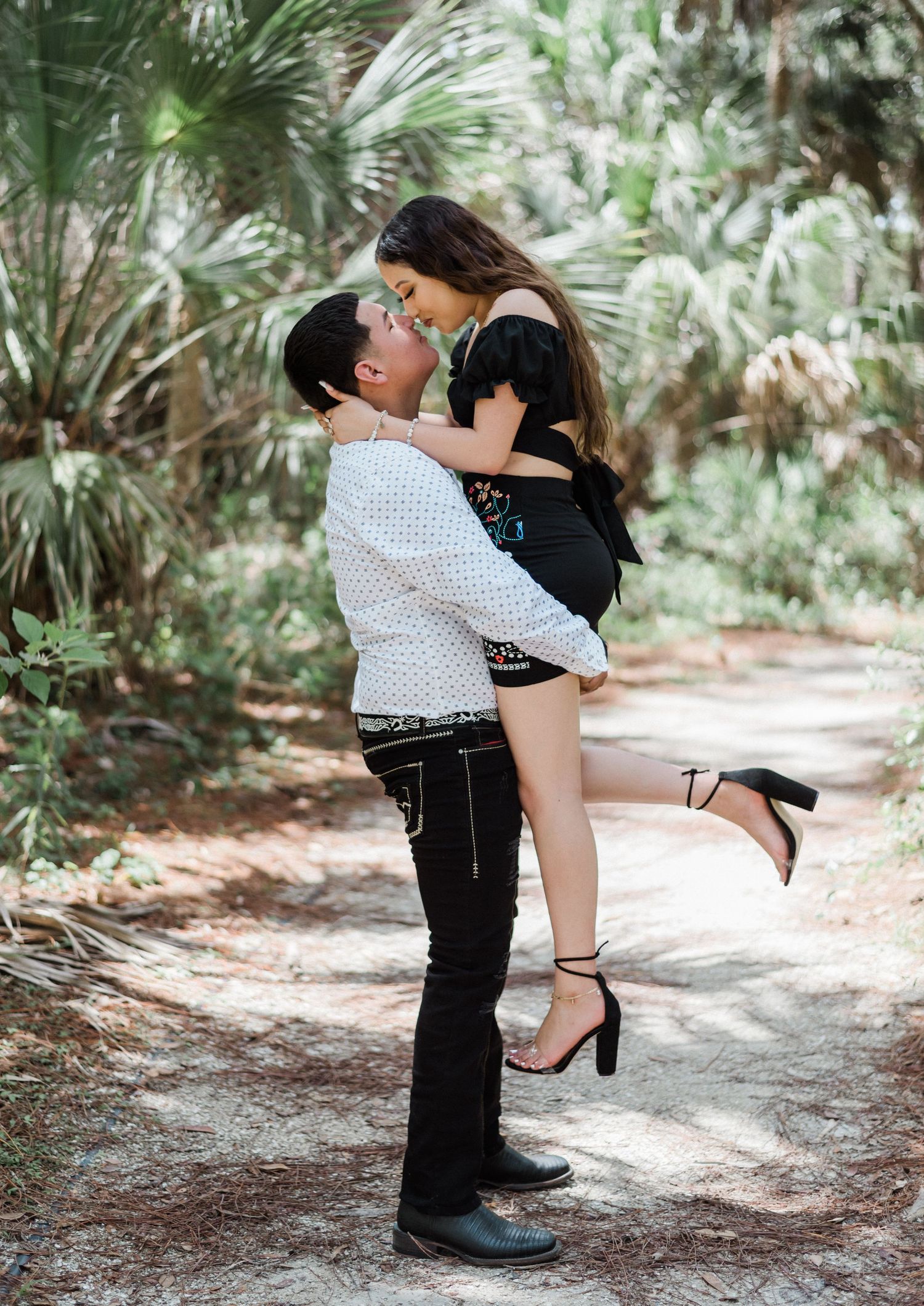 A romantic couple embraces on a sunlit forest path surrounded by tropical palm trees and greenery.