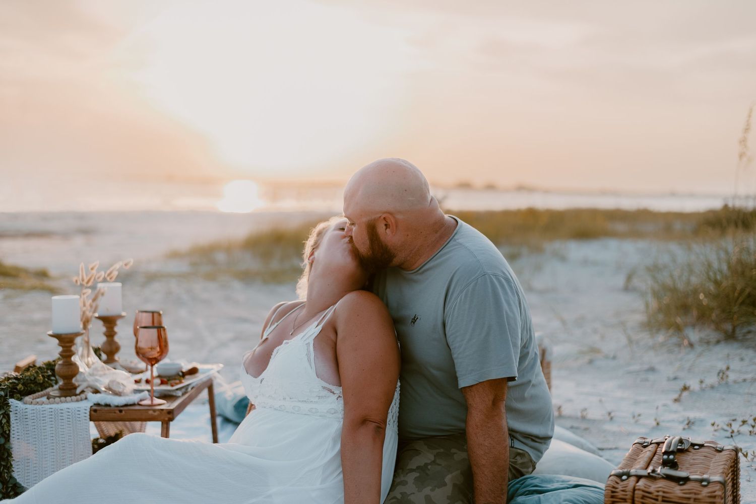 A romantic beach picnic setup at sunset with wine glasses and a cozy blanket on sandy dunes.