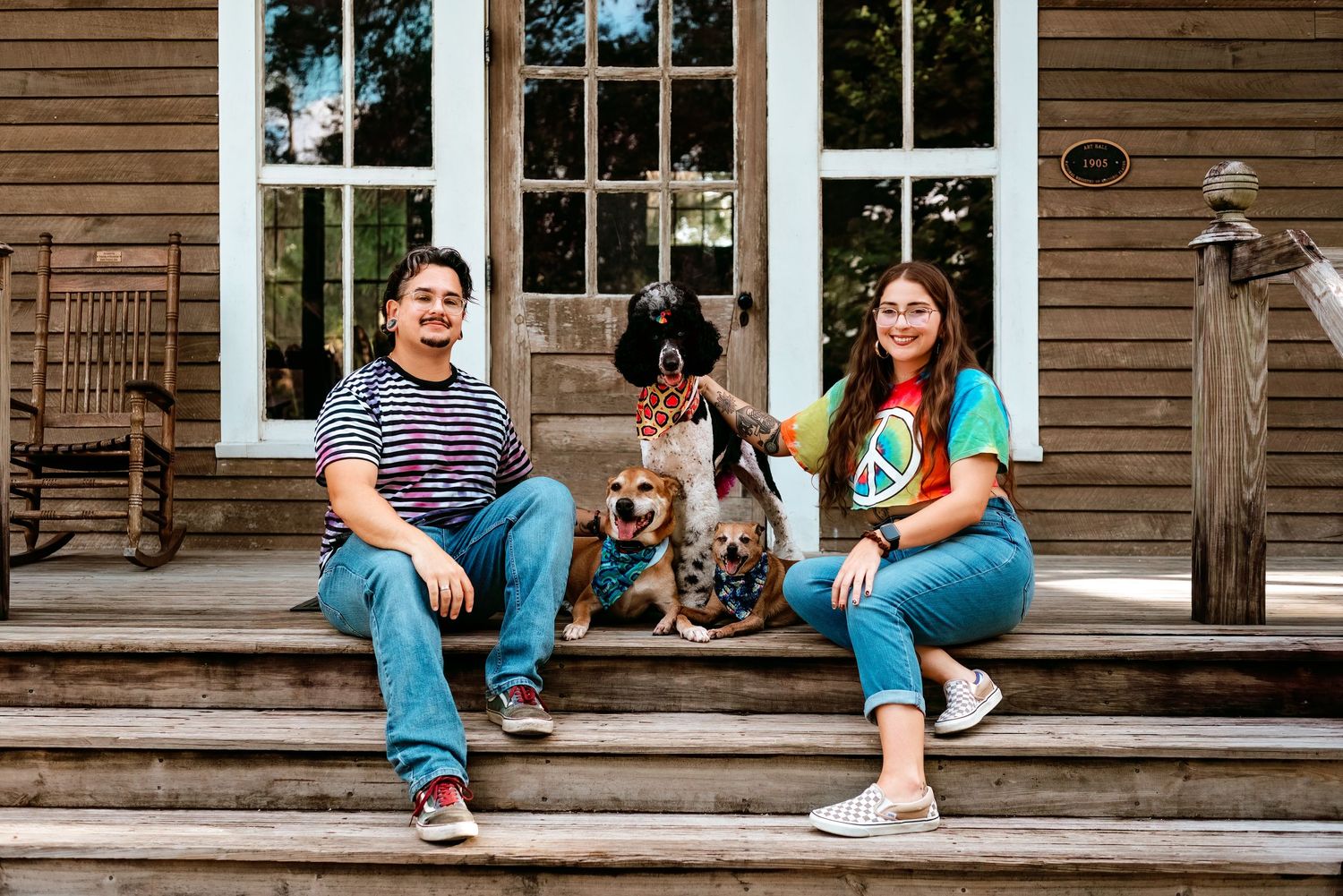 People relaxing on wooden porch steps with a dog at a rustic house with large windows.