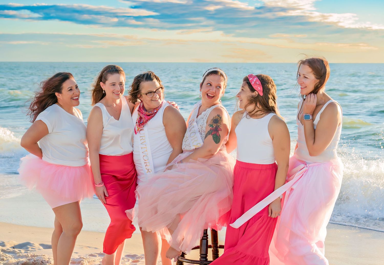 A group in pink and white dresses laughs and dances together on a sunny beach at sunset.