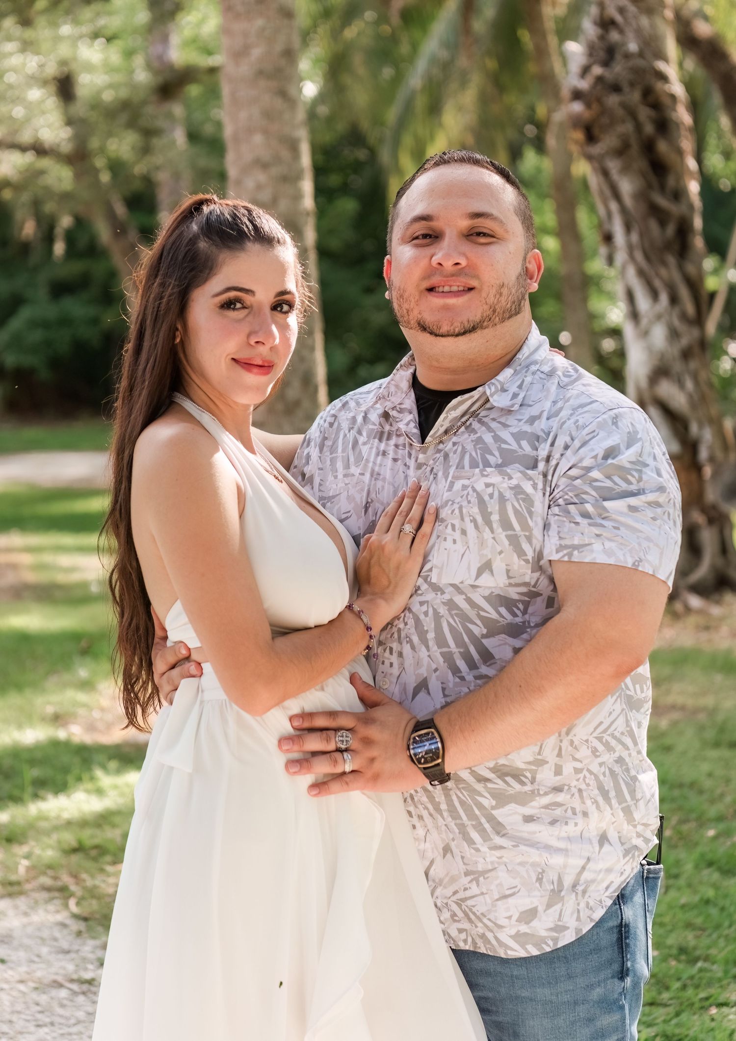 A couple in coordinating white and patterned outfits embraces while posing outdoors in a natural setting during a photo shoot.