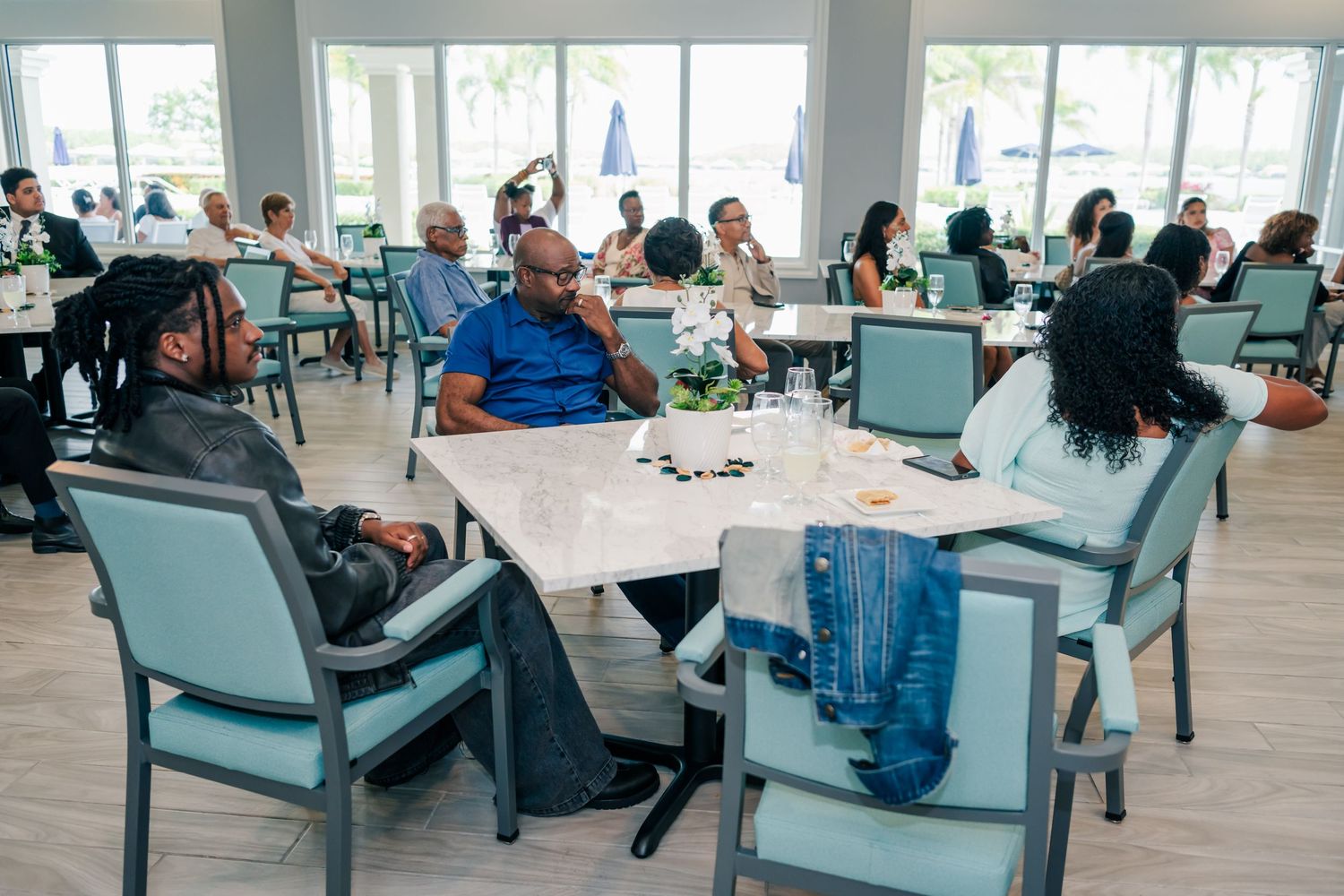 Guests networking at a bright, modern banquet hall with white tables and teal chairs during a social event.
