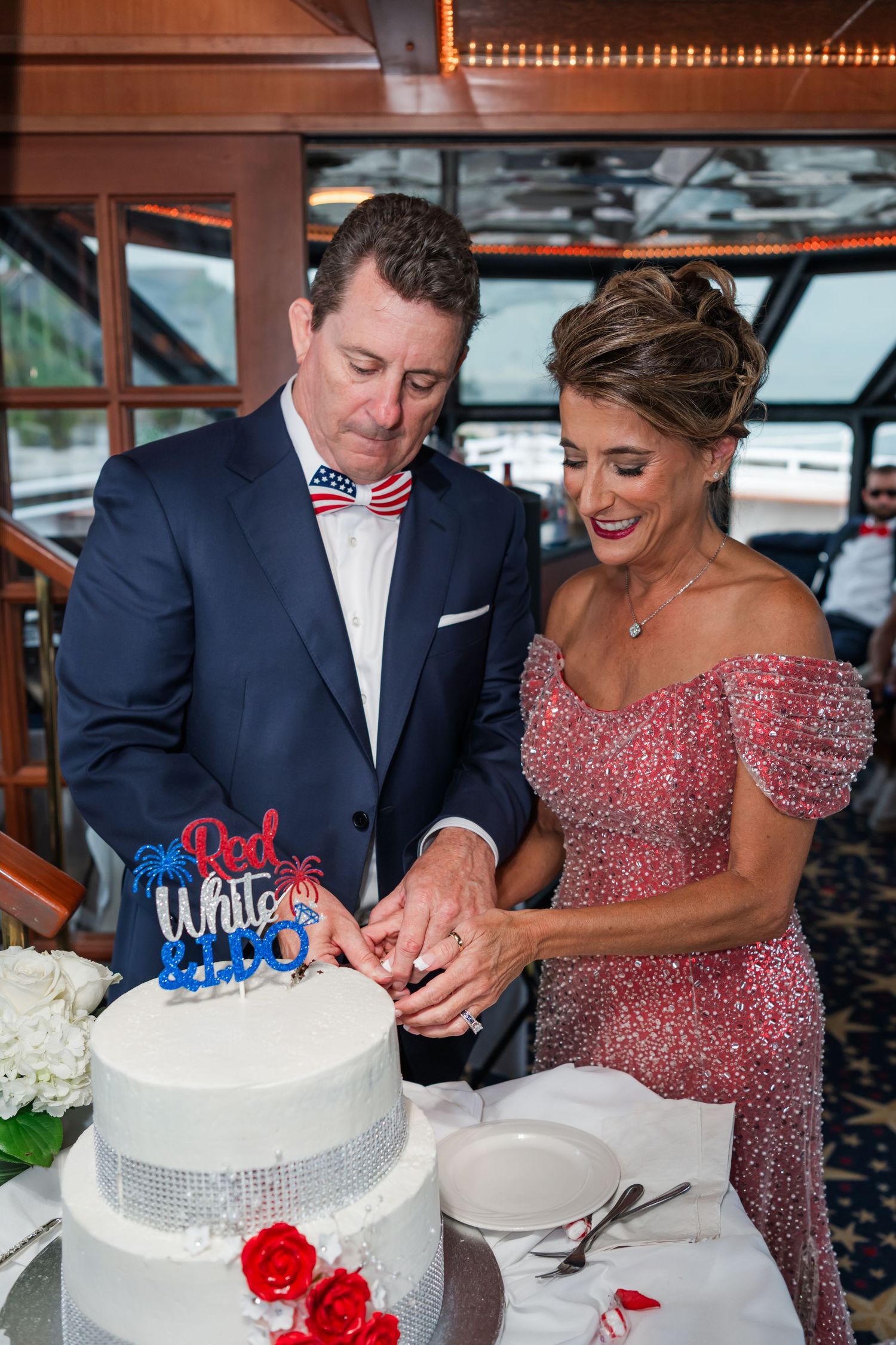Couple cutting a patriotic red, white and blue wedding cake on a boat, groom in navy suit, bride in pink sequin gown.