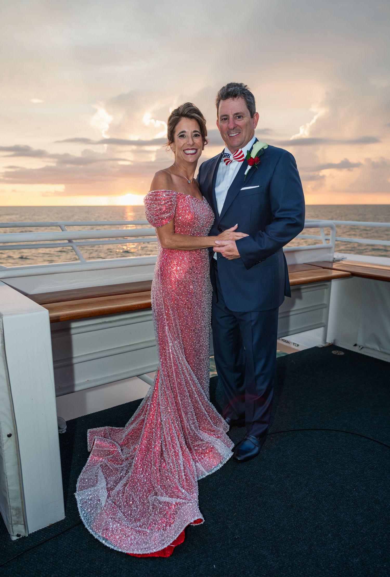Couple in formal attire poses on cruise ship deck at sunset, woman in pink sequin gown, man in navy suit.