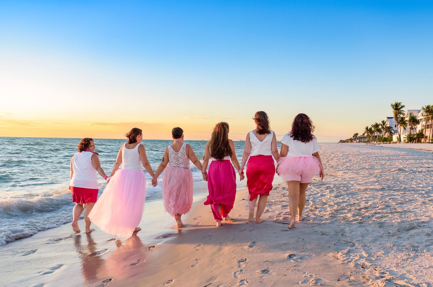 Group wearing pink and red dresses walking along a white sandy beach at sunset.