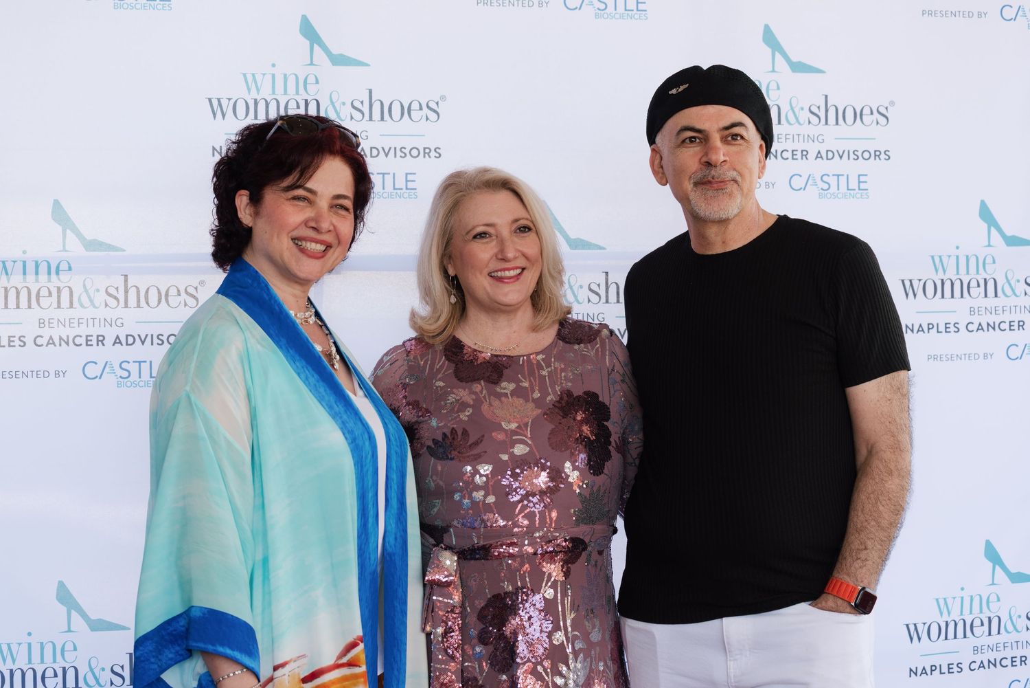 Three people pose together at a formal event with a branded backdrop featuring repeating logos.