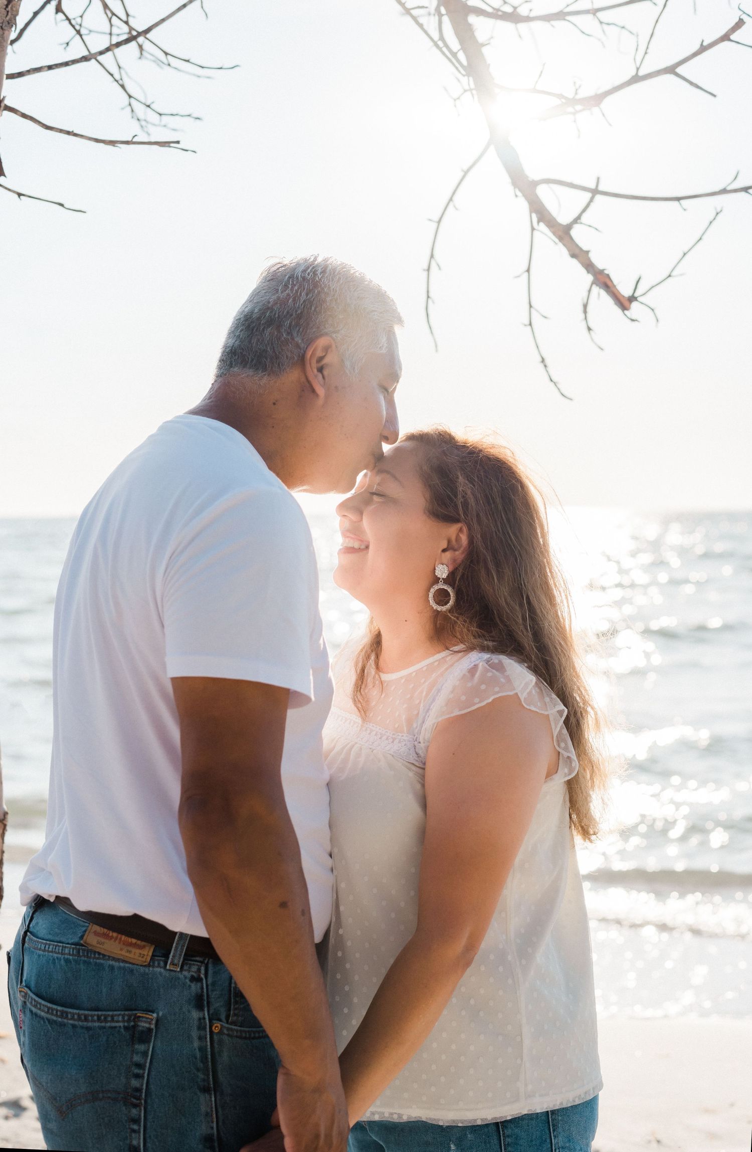 Couple sharing intimate moment on sunny beach with sparkling ocean waves and tree branches in background.