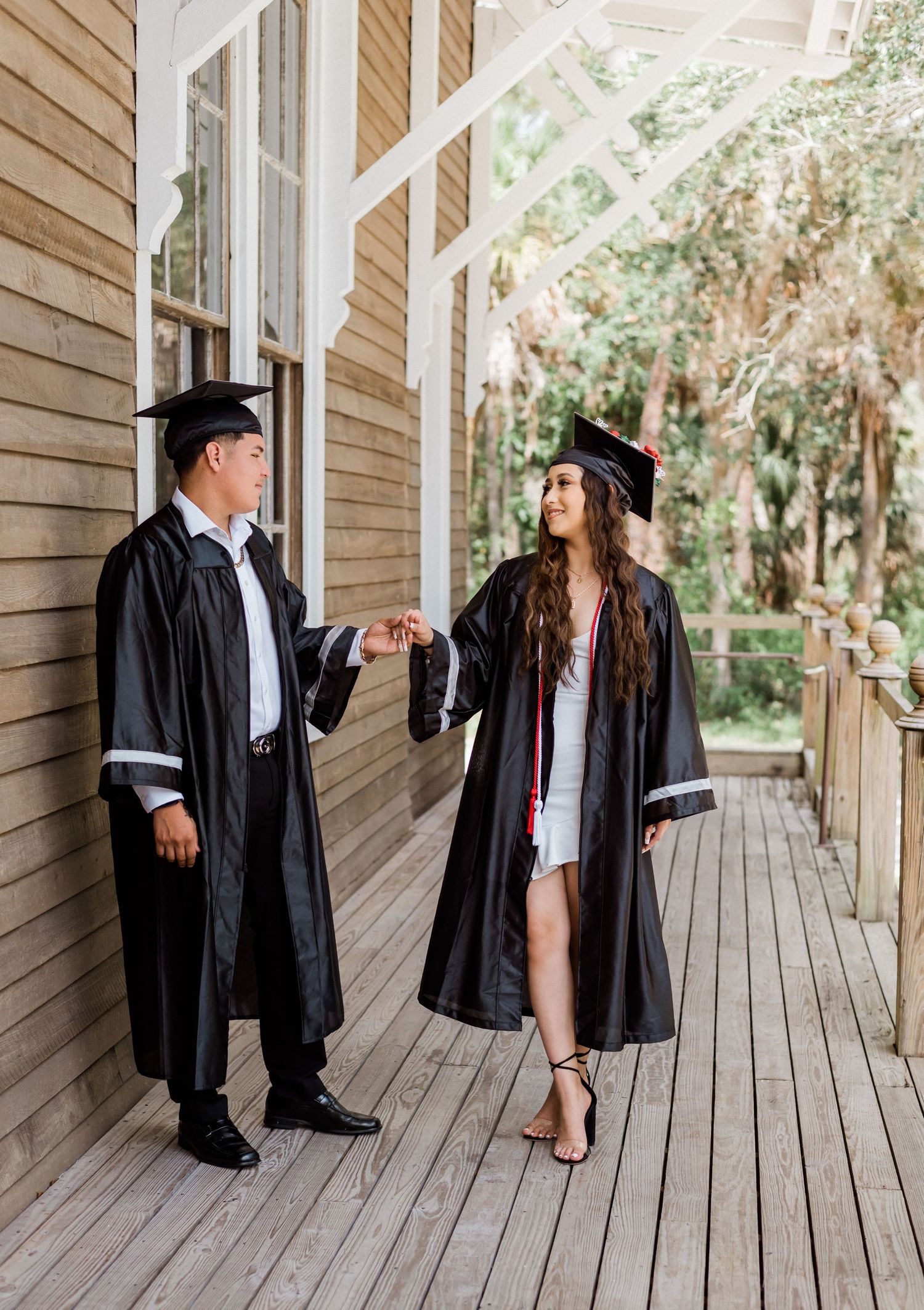 Two graduates in black caps and gowns standing on a wooden porch looking at each other during graduation celebration.