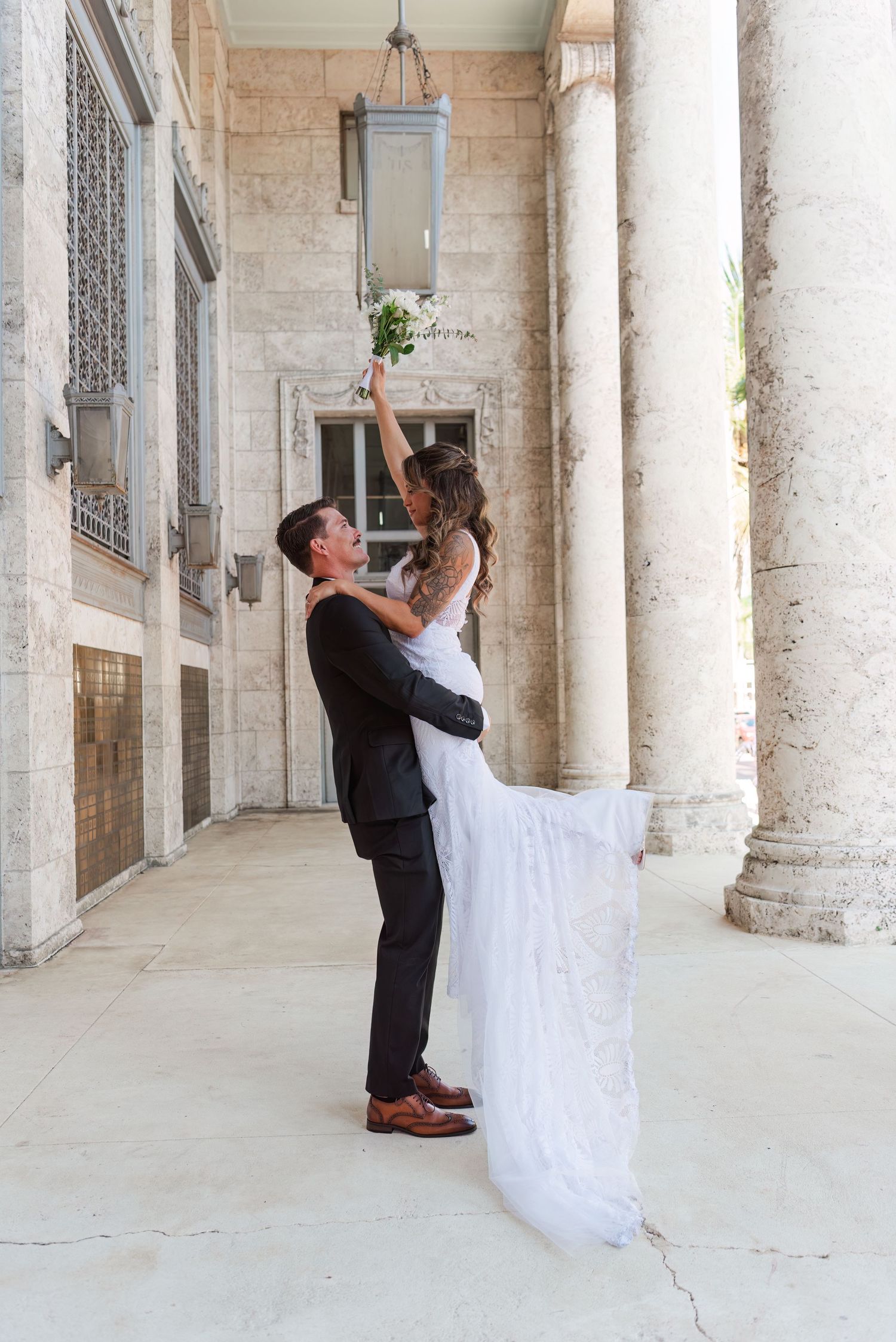 Groom lifting bride in white lace gown outside elegant stone building with columns as she raises bouquet.