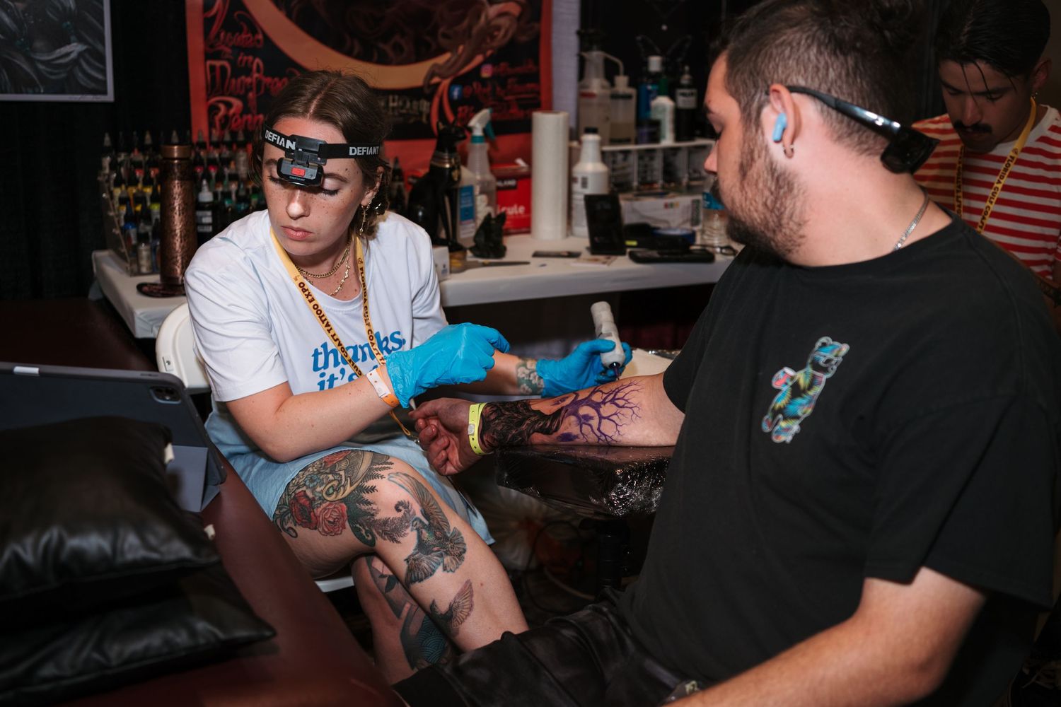 Tattoo artist wearing headlamp applies ink to client's arm at a tattoo convention.