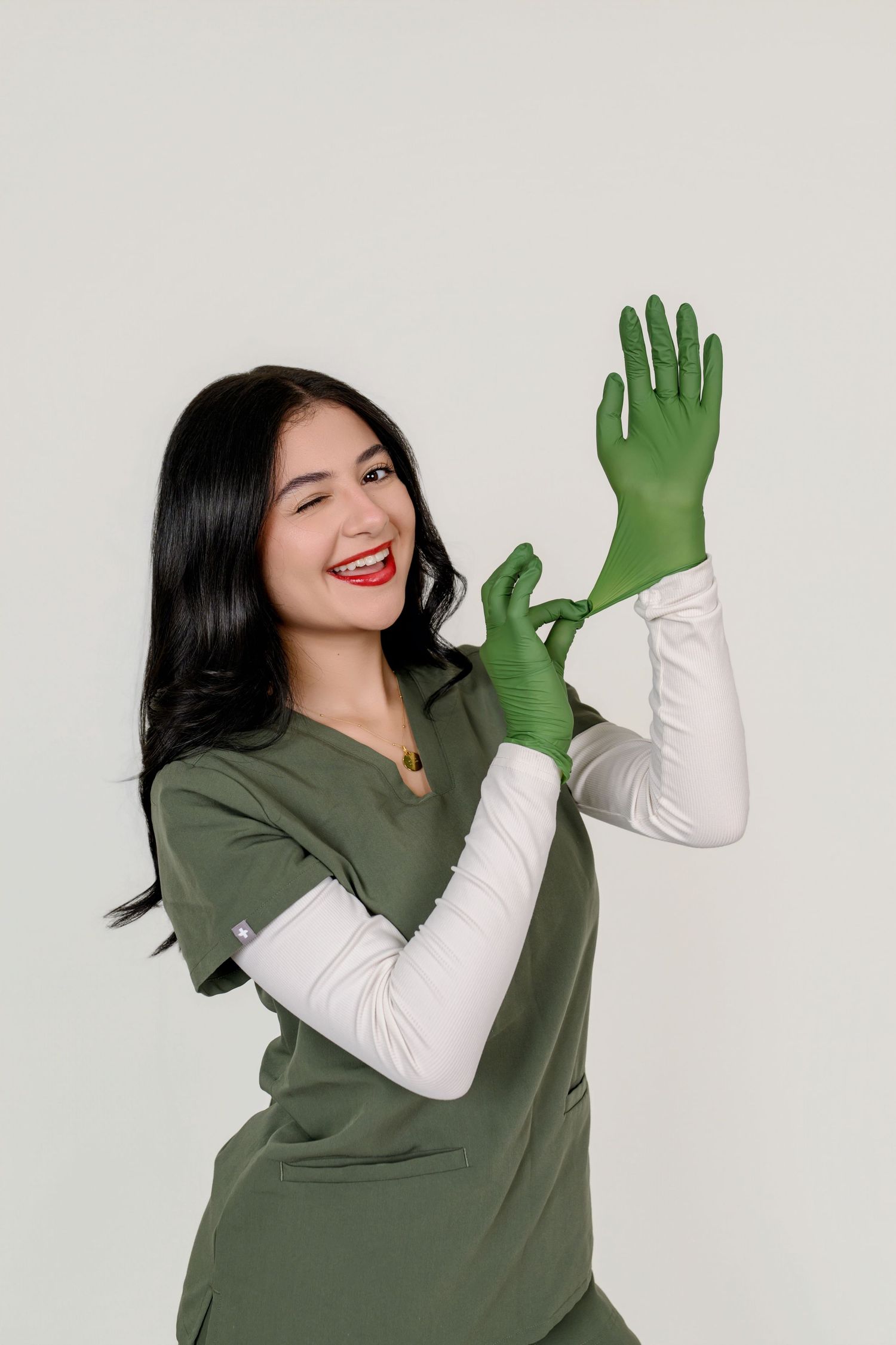 Smiling female healthcare worker in green scrubs putting on matching green latex gloves against white background.