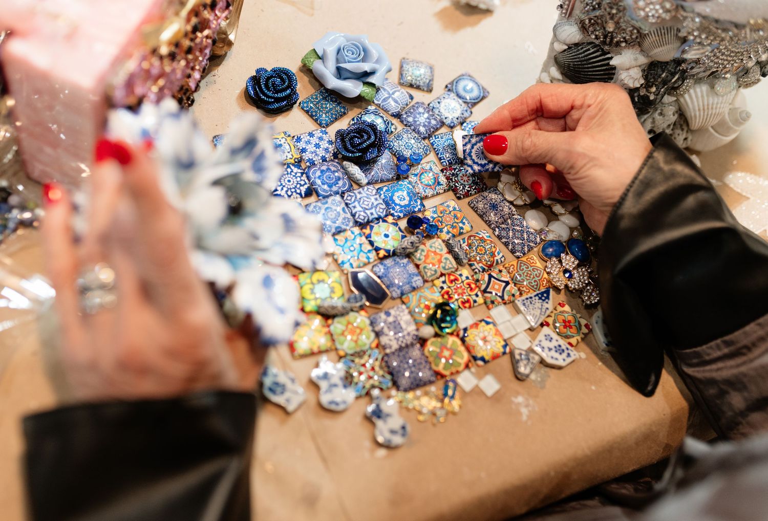 Hands arranging colorful blue patterned mosaic tiles and floral decorations on brown craft paper table.