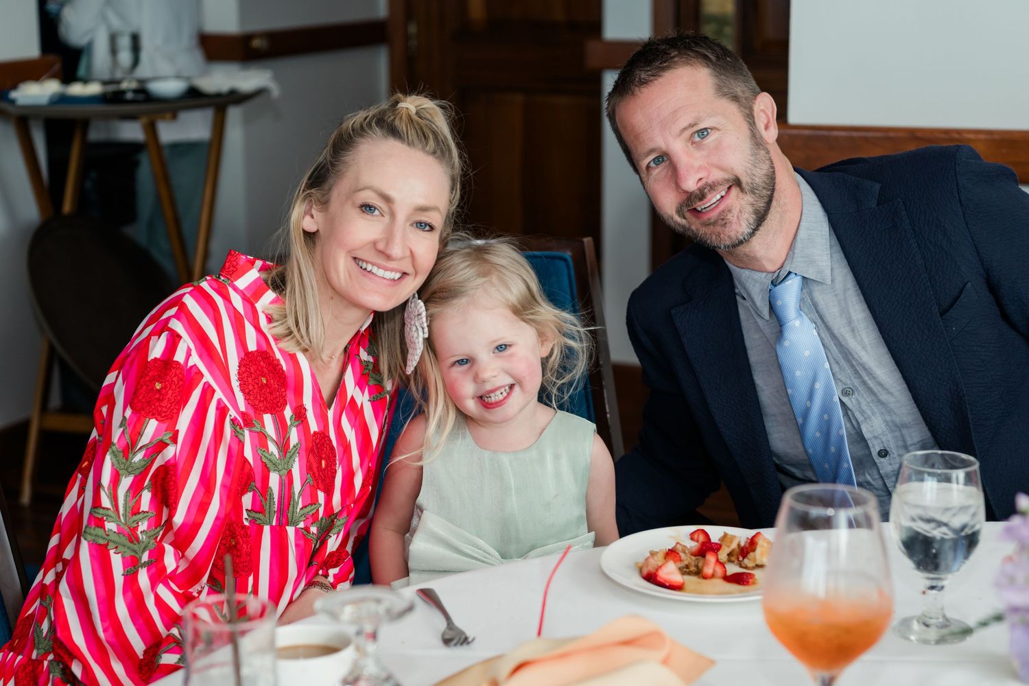 Family of three smiling at restaurant table with food and drinks, woman in red floral dress, young girl, man in suit.