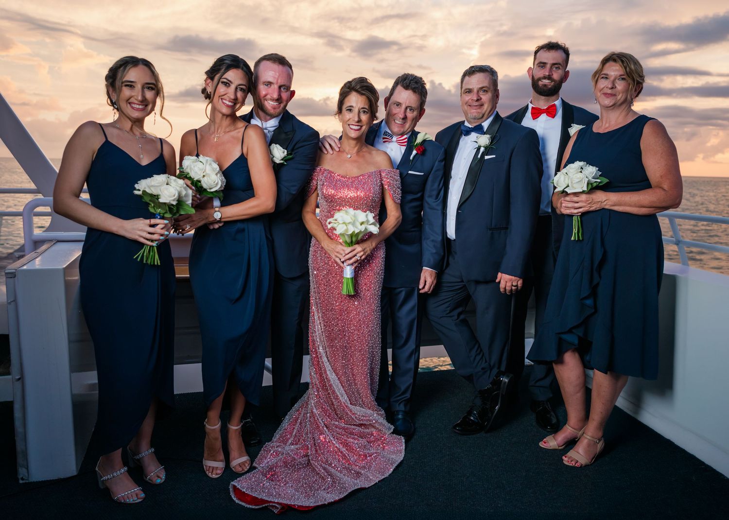 Wedding party posing on a boat deck at sunset, bridesmaids in navy dresses, groomsmen in suits, bride in pink sequin gown.