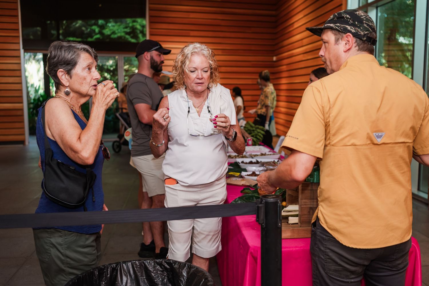 People gathered at an outdoor event, conversing near a pink-draped table with food and drinks on display.