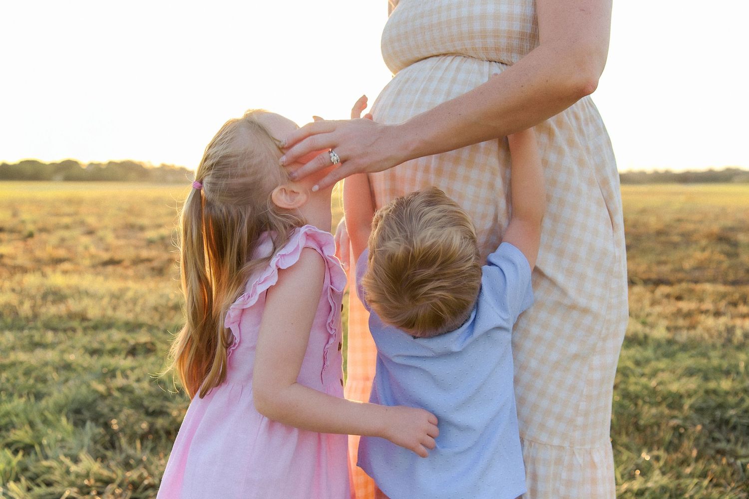 A family embraces during a golden sunset photoshoot in a grassy field wearing soft pastel colors.
