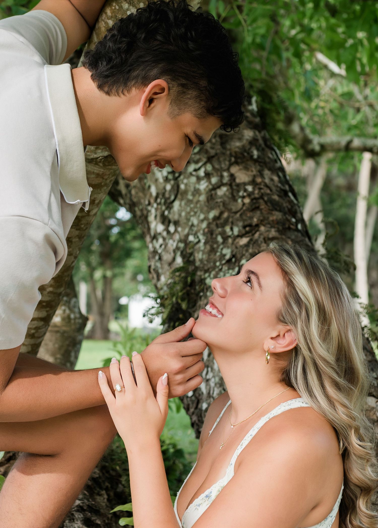 A couple shares a romantic moment under a tree, smiling at each other in a lush green outdoor setting.